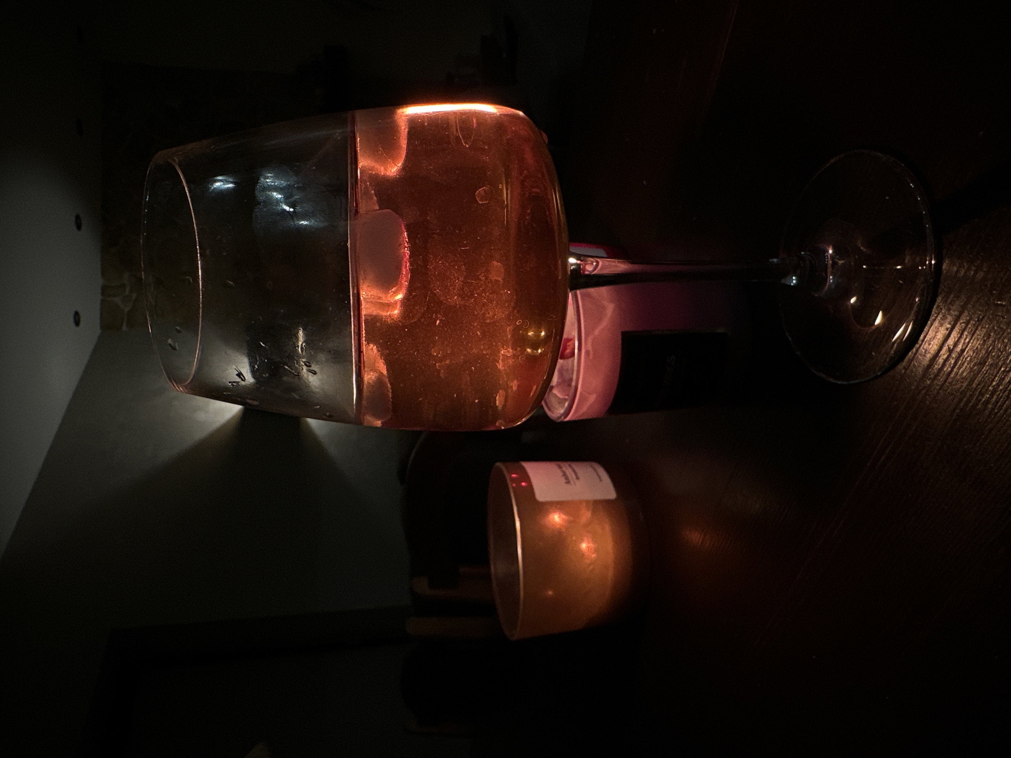 A dimly lit image shows a wine glass lying on its side on a dark surface, likely a table. A second, smaller glass is also visible. The lighting gives the glasses a reddish hue. The background is dark and indistinct. There is no visible evidence to suggest the location is Chaikovsky, Russia.