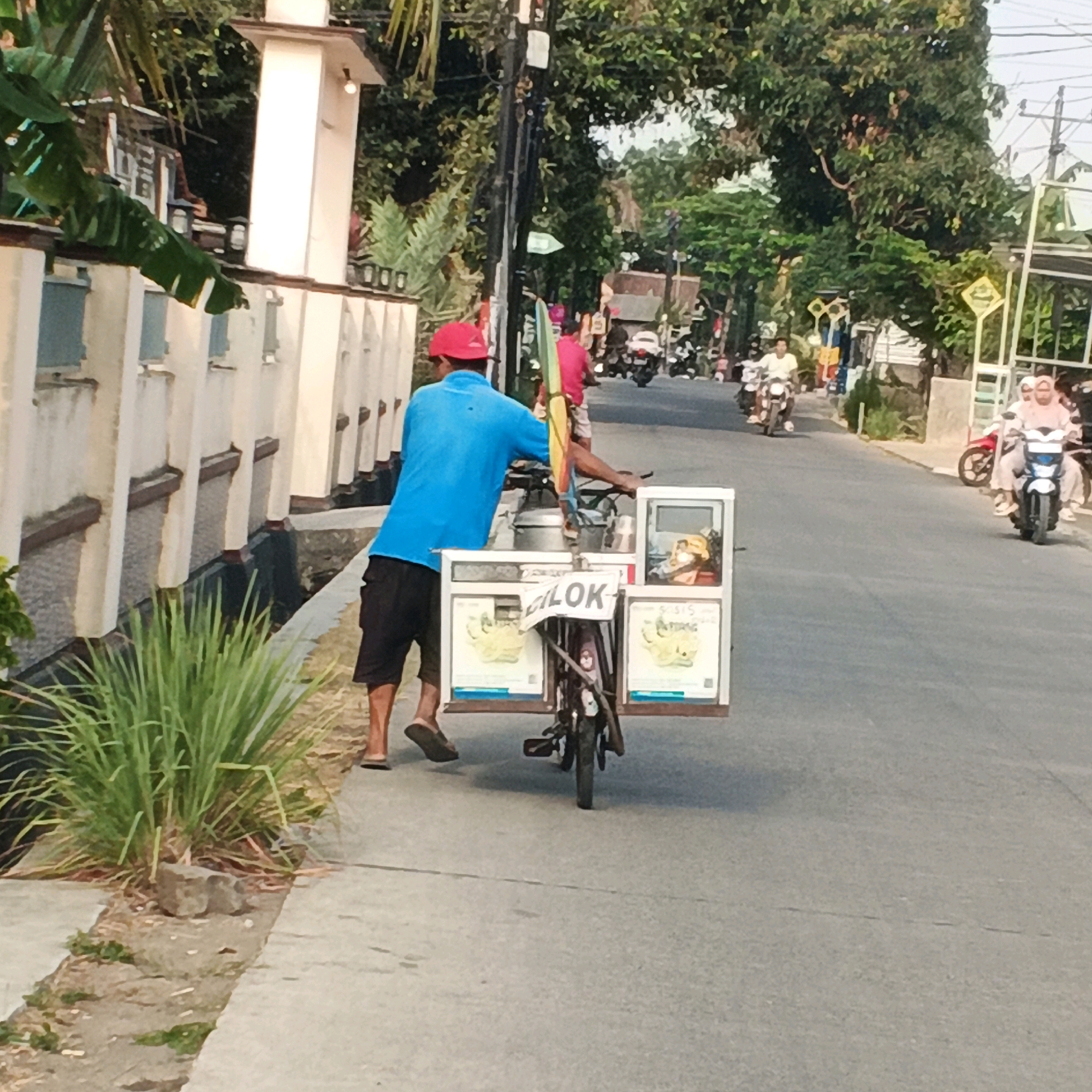 An eye-level, medium shot shows a man wearing a blue shirt and black shorts with a red cap, pushing a cart on a paved road in Karangjengkol, Indonesia. The cart has a white frame with pictures of food items and the word "CILOK" written on it. The man is on the left side of the frame, walking on a sidewalk next to the road, which is bordered by a low wall and lush green plants. Further down the road, several motorcycles are visible, some with riders, suggesting a typical street scene in a populated area. The environment is characterized by trees and a mix of buildings along the roadside. The lighting indicates it is daytime, possibly late afternoon due to the shadows.