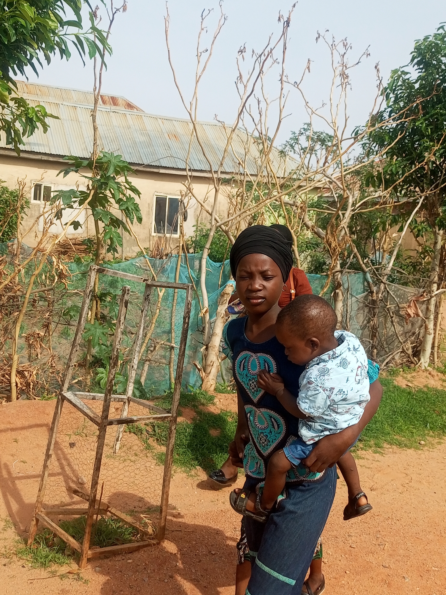 A young Black woman wearing a black hijab and a dark blue dress with a teal and white pattern is carrying a baby on her back. The baby is wearing a light blue shirt with a white pattern and denim shorts. They are standing in a dirt area next to a building with a corrugated metal roof. In the foreground, there is a wooden structure with chicken wire. There are trees and greenery in the background, and the sky is visible above. The location is stated to be Tempure, Nigeria.