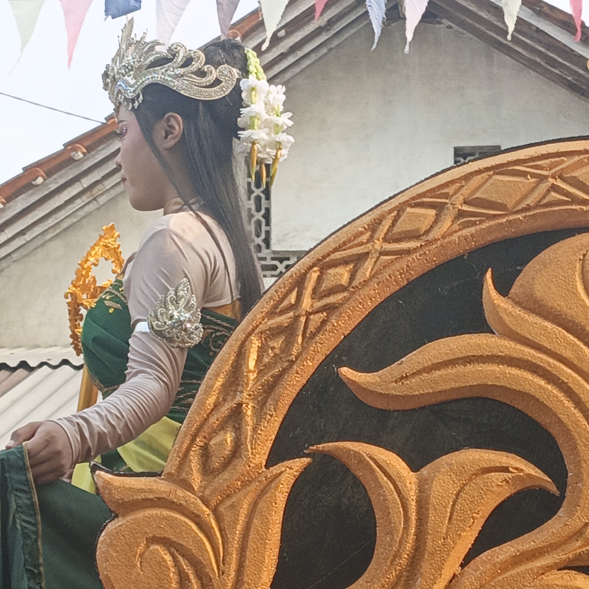 A person, adorned with a golden, intricate headdress and a white floral garland, is seated on a decorative float. They wear a cream-colored, long-sleeved garment and a green dress with gold trim. Their left arm rests on a green fabric, and a metallic, ornate cuff is visible on their right forearm. In the background, a building with a tiled roof and decorative flags are visible. The scene appears to be a parade or festival in Tanahbaya, Indonesia.