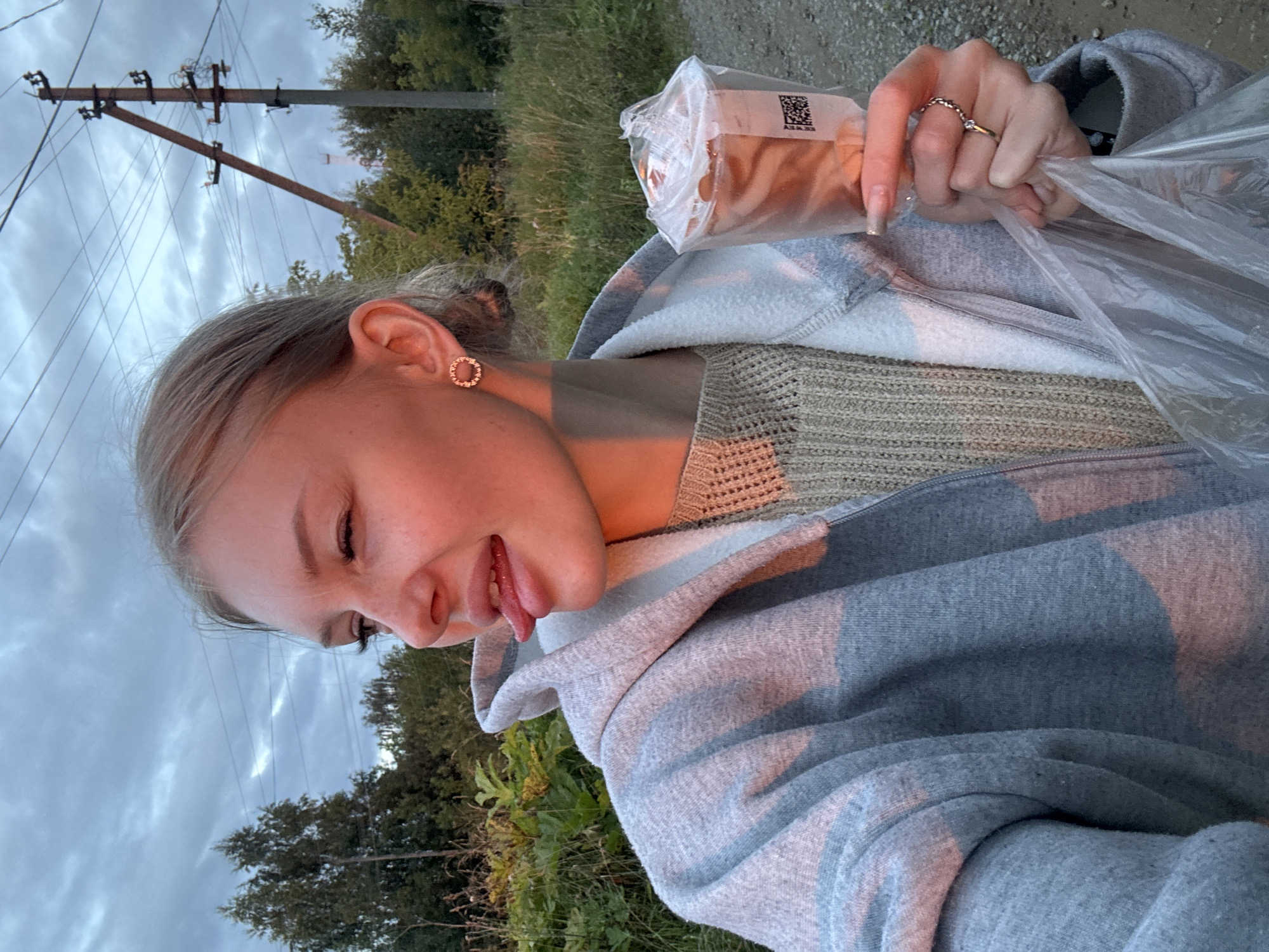 A young woman with blonde hair tied back is taking a selfie outdoors at dusk. She is wearing a grey hooded jacket over a beige knitted sweater. Her tongue is sticking out slightly to her right. She is holding a clear plastic bag containing a food item, possibly pastries, in her right hand. A ring is visible on her right ring finger. The background shows trees, vegetation, a dirt road, and a utility pole with power lines. The lighting suggests the sun is low on the horizon, casting a warm glow on her face. The location is identified as Borodulino, Russia.