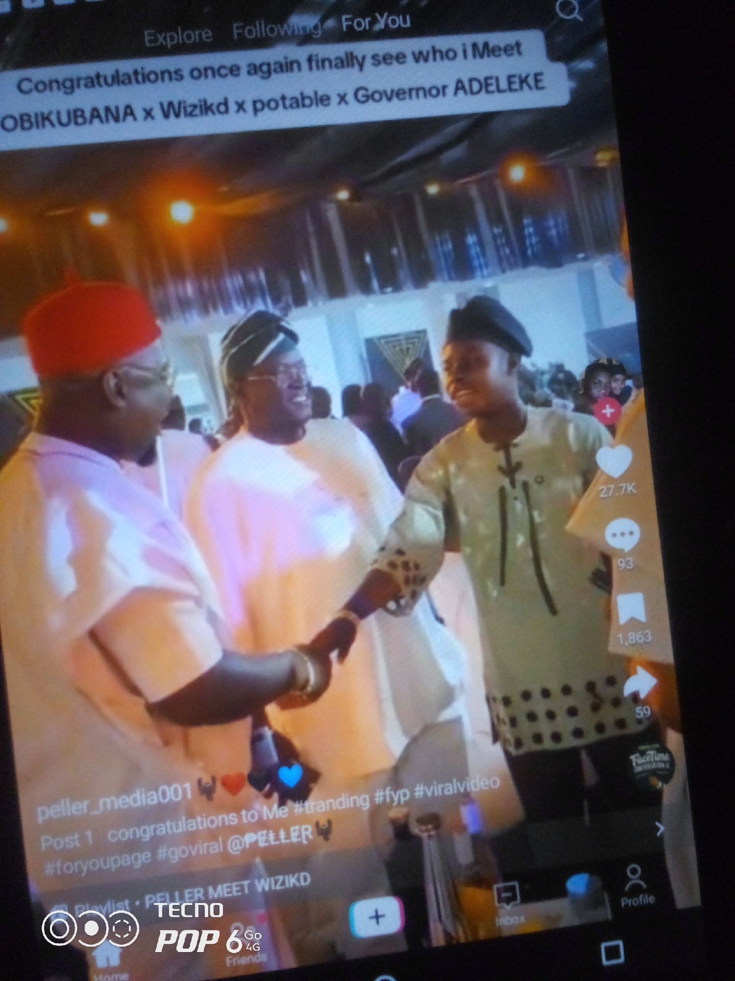 The image captures a screenshot of a video playing on a mobile device, likely a TikTok video given the UI elements like like counts, comment counts, and share buttons. The video displays a group of people, presumably in Nigeria, given the mention of "Governor ADELEKE" which is a Nigerian politician.
In the foreground, three men are the primary subjects. The man on the left wears a red cap and a light-colored traditional Nigerian attire, possibly an agbada. He is looking to his right and appears to be interacting with the man next to him. The central figure is also in light-colored traditional attire and a headwrap, and he is smiling and looking towards the left, seemingly engaged in conversation. To the right, a younger man is dressed in a dark green traditional outfit with intricate black patterns on the sleeves and a black cap. He is looking to his left and appears to be in motion or speaking.
The background suggests an indoor event, possibly a celebration or gathering, with warm lighting and blurred figures of other attendees. Some of the UI elements on the screen include text overlays such as "Congratulations once again finally see who i Meet OBIKUBANA x Wizikd x potable x Governor ADELEKE", and hashtags like "#trending #fyp #viralvideo". Further down, there is text related to a profile handle "peller_media001" and a playlist entry "PELLER MEET WIZIKD". The bottom of the screen shows interface elements for a mobile application, including icons for home, inbox, and profile, along with the branding "TECNO POP 6 4G".
The location within the city of Howayi, Nigeria, cannot be determined from the visual information provided in the image. The image is a digital screenshot and lacks any geographical markers or context that would pinpoint its precise location within Howayi or any other city in Nigeria.