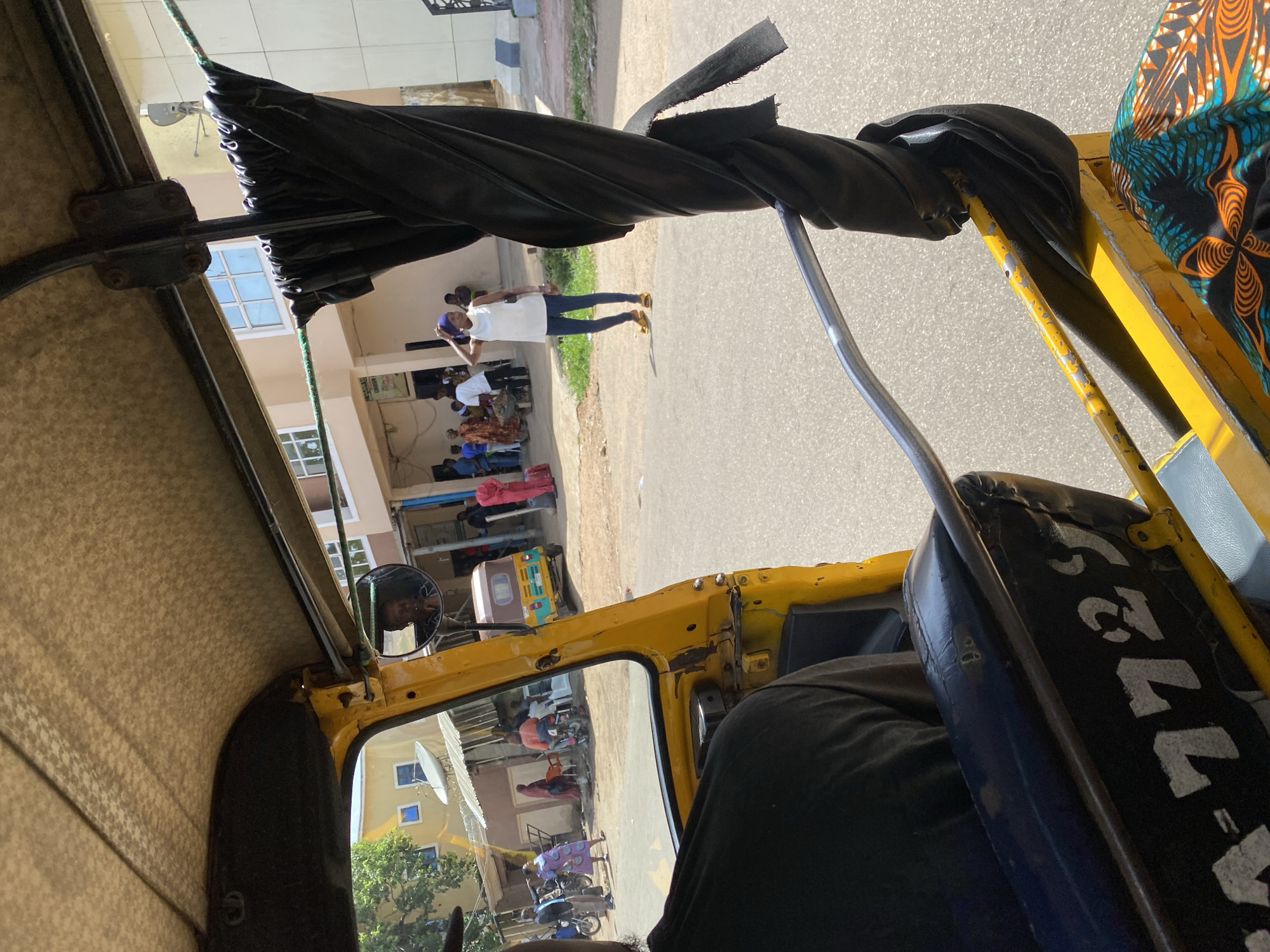 The image, taken from inside a yellow autorickshaw in Jos, Nigeria, captures a street scene. The foreground shows the interior of the autorickshaw, with a black seat bearing the registration number "A-7729" and a colorful patterned fabric. A person's back, wearing a black shirt, is visible in the driver's seat. The view through the windshield reveals a street with a few people and vehicles. In the midground, several individuals are seen near buildings. One woman in a white top and blue jeans, wearing a blue headscarf, stands with her hand on her head. Another person in a pink outfit stands further down the street. A yellow and brown autorickshaw is parked on the road, and a motorcycle is also visible. The background consists of a building with windows and a satellite dish. The overall environment appears to be a moderately busy urban street.