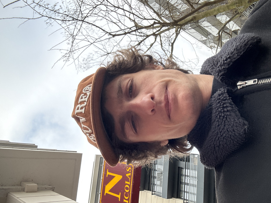 A male individual is captured in an upward-angled, close-up photograph, standing outdoors on an overcast day. He is positioned in front of a commercial and residential streetscape at 10 Rue de Paris, Joinville-le-Pont, France. The subject has medium-length, wavy brown hair and is wearing a brown corduroy baseball cap with the word "OUVREA" embroidered in white on the front. He is also dressed in a black jacket featuring a prominent black faux fur or fleece collar and a visible silver-toned zipper down the front. His expression is neutral as he looks directly at the camera.
In the background, to the subject's left, a segment of a red commercial sign displaying "N NICOLAS" in large yellow capital letters is clearly visible, identifying a Nicolas wine shop. Behind and below this sign, a multi-story modern building with dark-framed windows and dark-railed balconies is partially visible. To the subject's right, bare branches of a deciduous tree extend upwards against a bright, cloudy sky, indicating a winter or early spring season. Another building with a reflective facade is faintly discernible behind the tree branches.