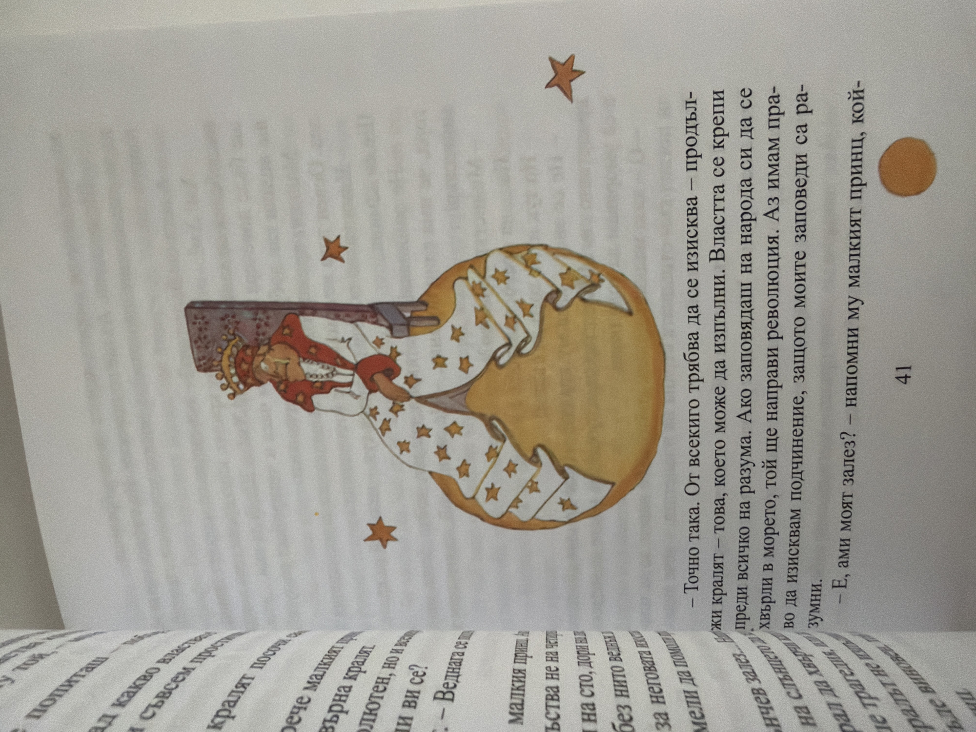The image shows an open book with text and an illustration. The illustration is of a king with a crown, sitting on a globe. He is wearing a regal robe decorated with stars. There are also stars scattered around the globe. The text is in Bulgarian. The left page has text at an angle, indicating it's from the edge of a book. The right page has a picture and text in straight lines. One snippet of text reads "– Точно така. От всекиго трябва да се изисква – продъл- жи кралят – това, което може да изпълни. Властта се крепи преди всичко на разума. Ако заповядаш на народа си да се хвърли в морето, той ще направи революция. Аз имам пра- во да изисквам подчинение, защото моите заповеди са ра- зумни." Another snippet reads "– Е, ами моят залез? – напомни му малкият принц, кой- 41".
The image does not contain any information about the city of Pazardzhik, Bulgaria.