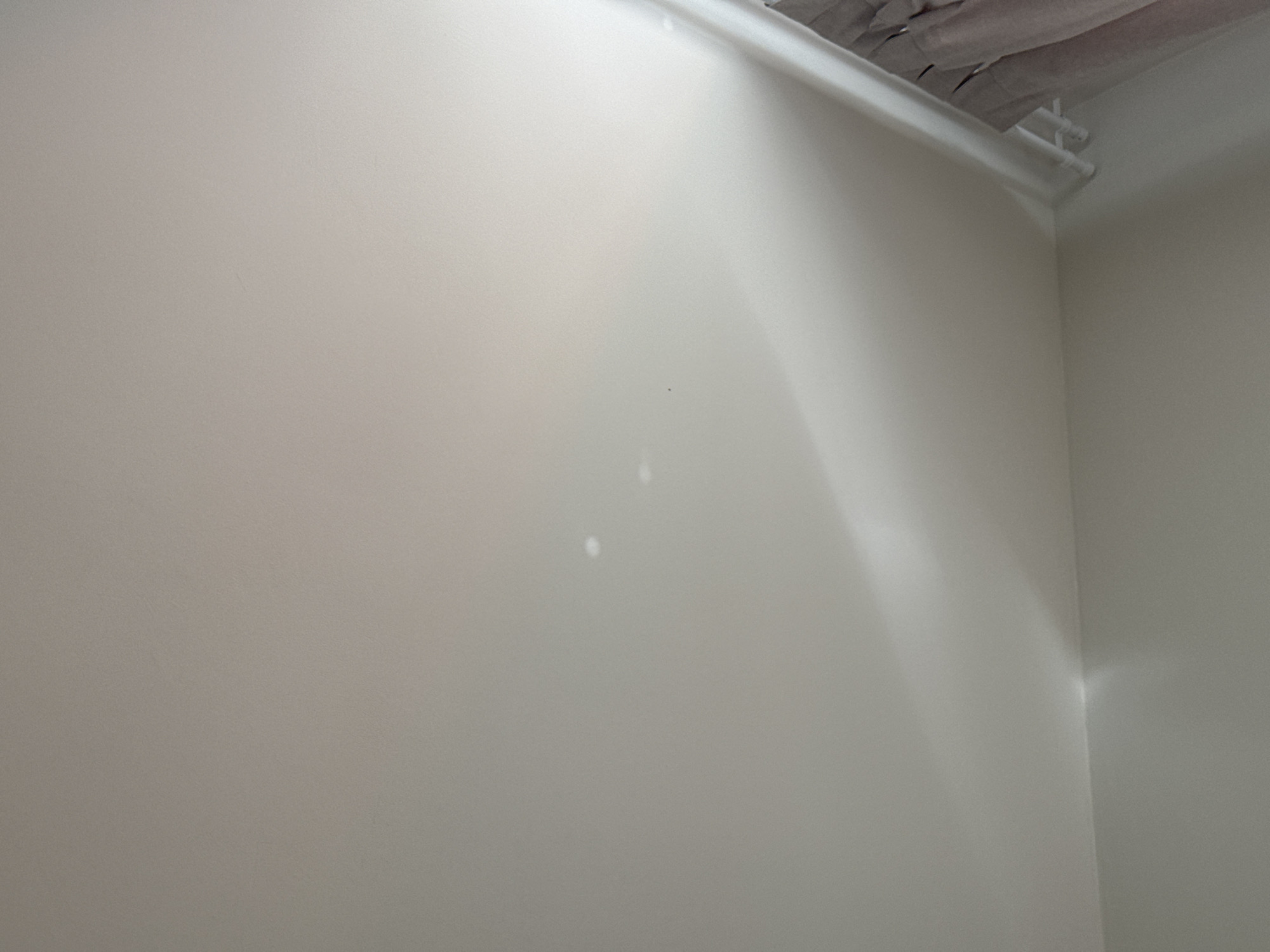 The image shows a corner of a room. The walls are painted in a light neutral color. A white pipe runs along the ceiling, near the corner. The ceiling above the pipe appears to be unfinished with visible material. There are some minor imperfections on the wall. No people are visible. The location cannot be confirmed as Люк, Russia, from the visual information provided.
