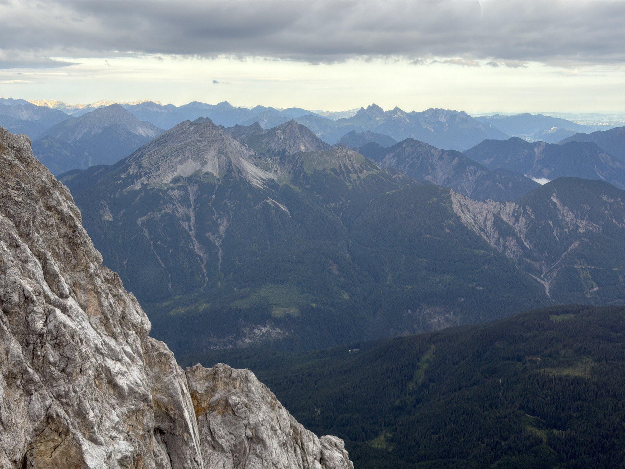 The image shows a panoramic view of a mountainous landscape from a high vantage point, likely a rocky peak in the Zugspitze massif near Grainau, Germany. In the foreground, a steep, weathered, grey rock face dominates the left side of the frame, showing signs of erosion with jagged edges and deep crevices. Beyond this, a vast expanse of rolling mountain ranges stretches into the distance. These mountains are covered in dark green coniferous forests in the middle ground, transitioning to lighter green and then a hazy blue in the far distance. Some of the peaks in the mid-distance show grey, rocky slopes. The sky is overcast with heavy, dark grey clouds in the upper portion, with lighter, broken clouds and patches of pale sky nearer the horizon, suggesting either early morning or late afternoon lighting. The overall atmosphere is one of natural grandeur and ruggedness, with no human figures or structures immediately visible in the primary view, though there appear to be very small, distant buildings nestled within the forest in the middle ground.