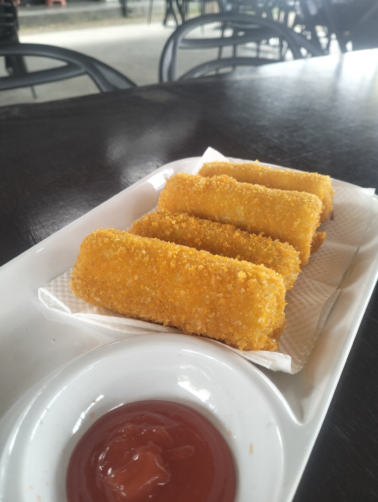 A plate of crispy fried spring rolls sits on a dark wooden table, accompanied by a small white dish filled with ketchup. The spring rolls are golden brown and coated in breadcrumbs, arranged in a neat row. The scene appears to be set at a cafe or restaurant, possibly in Lhokseumawe, Indonesia, given the ambient lighting and the style of furniture visible in the blurred background.