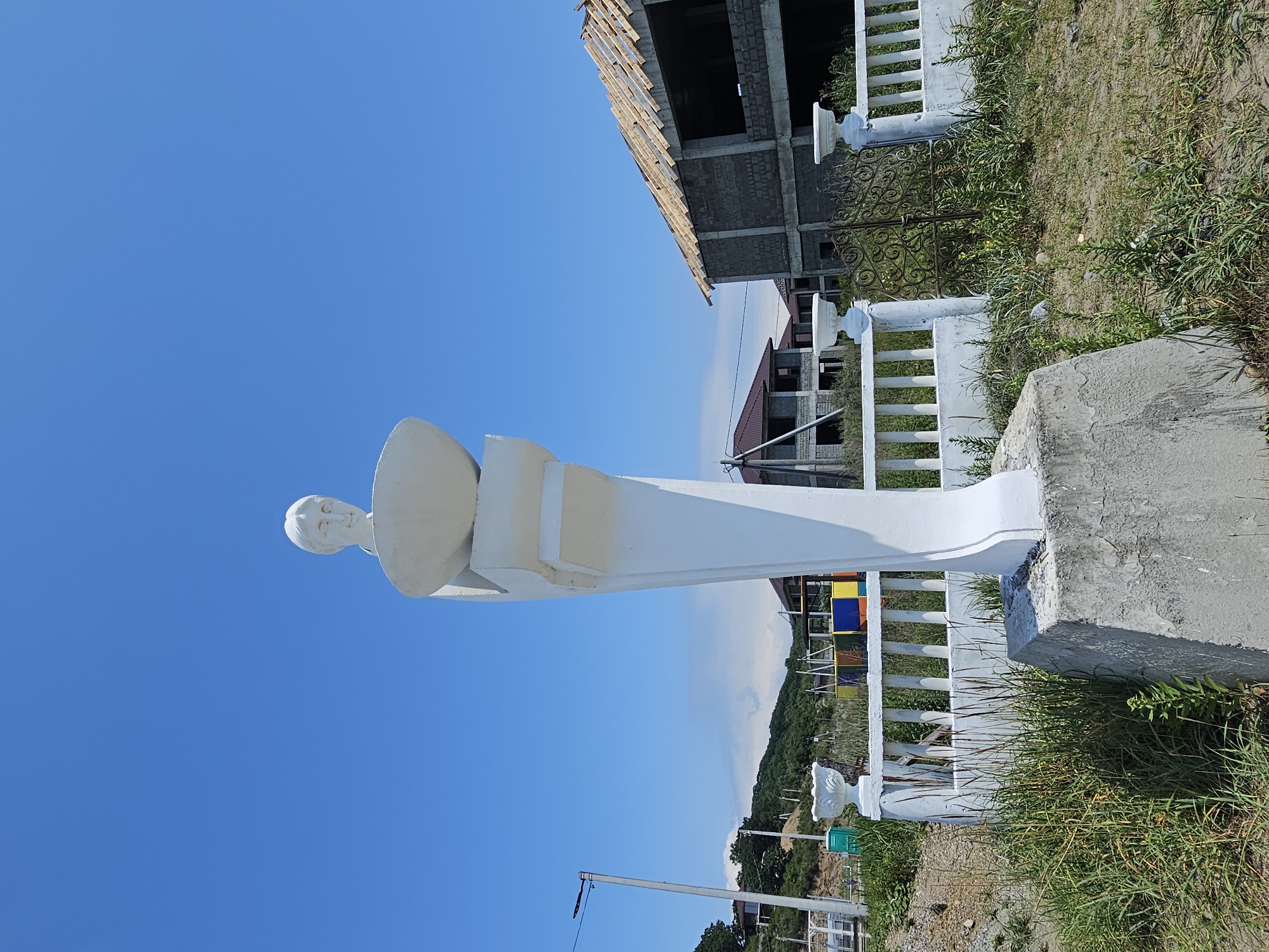 In Nakhodka, Russia, a white, classical-style bust of a person with short hair and a serene expression sits atop a pedestal in an outdoor setting. The bust is positioned centrally, slightly to the right of the frame. Behind the bust, a white, decorative fence runs horizontally, punctuated by white urns. Further in the background, the construction of several houses is evident, with visible concrete block walls and exposed wooden roof framing. A few trees and hills are visible on the horizon under a clear blue sky. The ground in the foreground is covered with dry grass and dirt.