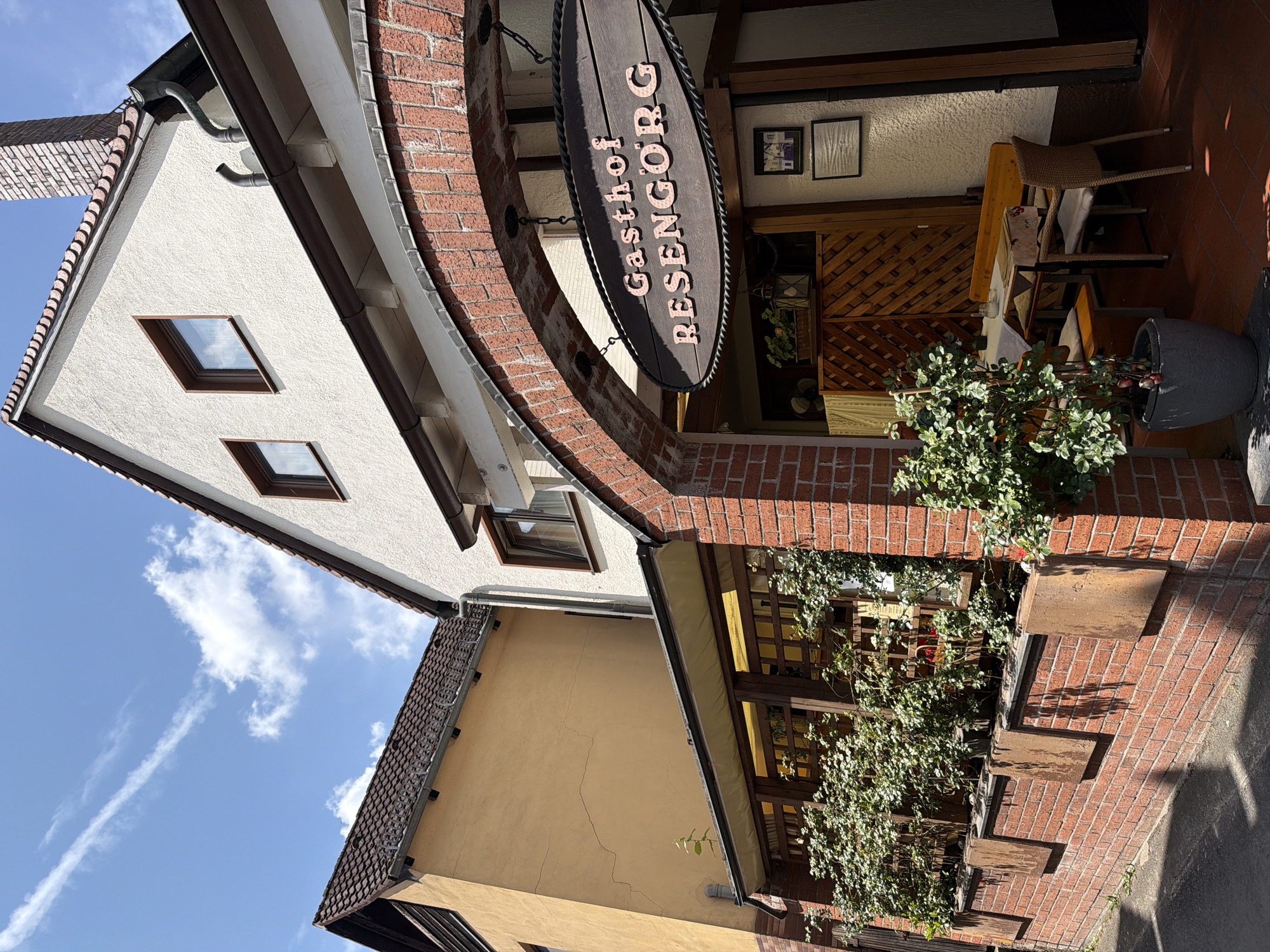 The image shows the exterior of a building, identified as "Gasthof RESENGÖRG" in Ebermannstadt, Germany. The building has a light-colored facade with a tiled roof and a prominent brick archway. Hanging from the archway is a dark wooden sign with the name of the establishment.
To the left of the main building, a yellow-colored building with a tiled roof is visible. The area beneath the archway appears to be an entrance to the establishment, possibly a restaurant or inn, with an outdoor seating area. This seating area includes wooden tables and chairs, covered by a white tablecloth, and surrounded by potted plants and greenery. The ground is paved with reddish tiles.
The scene is set on a sunny day, as indicated by the bright lighting and shadows cast on the ground. The sky is blue with scattered white clouds. The architecture suggests a traditional European style.