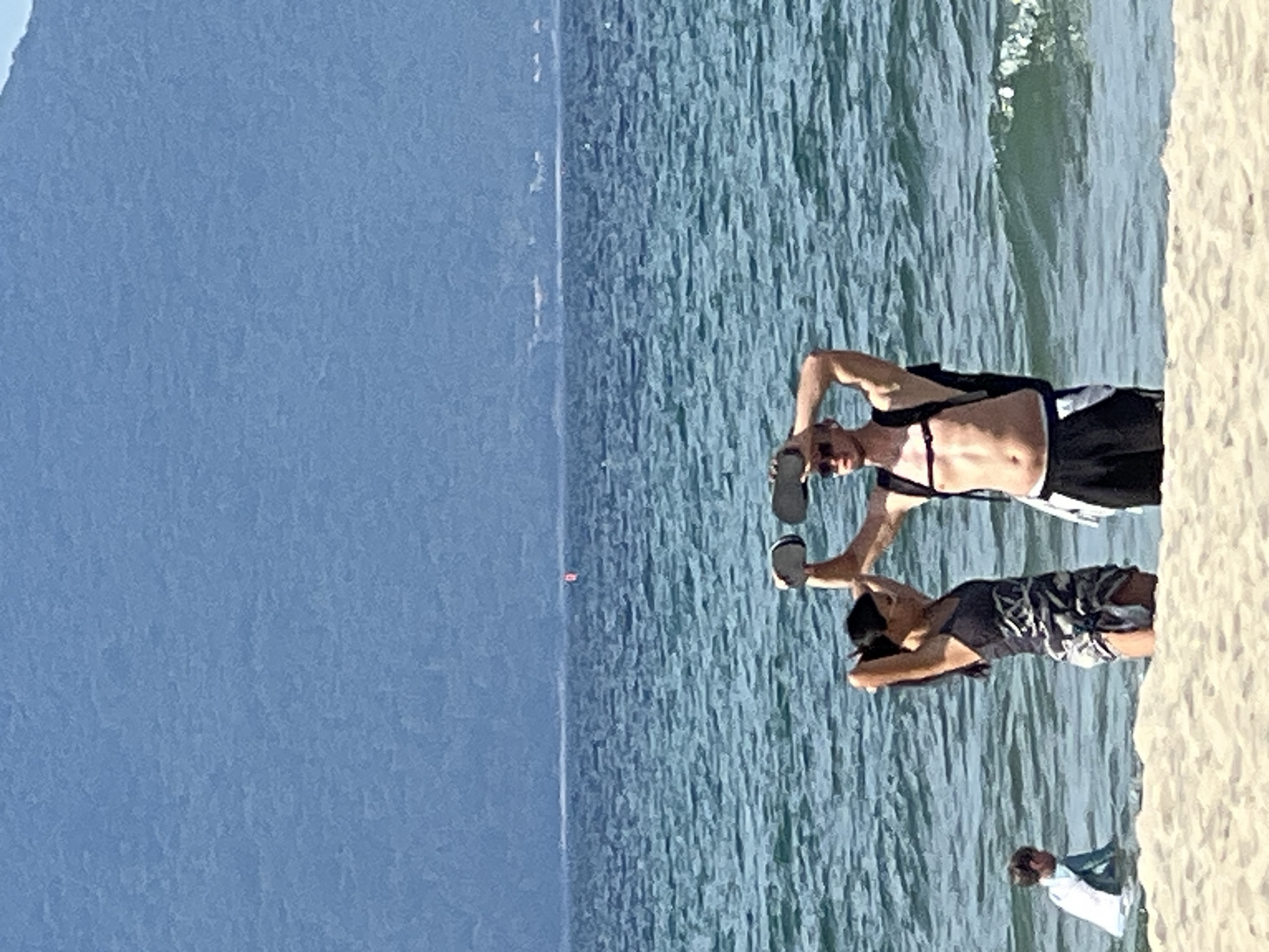 A candid, eye-level, outdoor, mid-shot photograph captures a beach scene on a sunny day in Đà Nẵng, Vietnam. In the foreground, the sandy beach is met by the turquoise ocean. A man and a woman stand in the shallow water. The man, wearing sunglasses, a black backpack, and black shorts with a white waistband, holds two round, white objects above his head. The woman, to his left, wears a dark-colored swimsuit with a camouflage pattern and is adjusting her hair. To the left of the couple, a young child in a white and blue rash guard sits in the water. In the background, a large, hazy mountain range with a blue tint dominates the horizon, separating the sea from the sky. The ocean is choppy, with small waves.