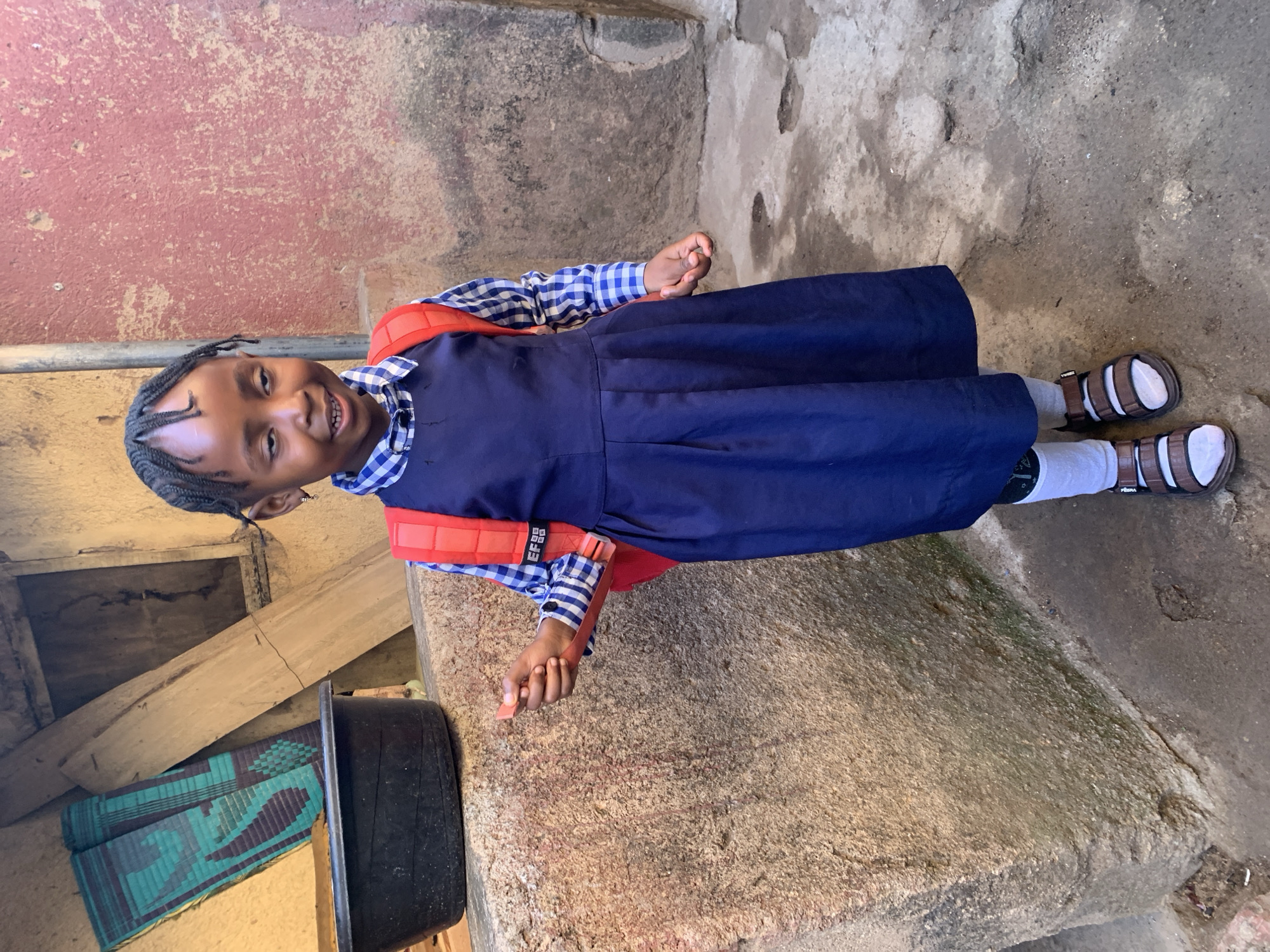 A young girl stands in front of a concrete wall, wearing a blue school dress, a blue and white checkered shirt, white socks, and brown sandals. She has a red backpack on. The location appears to be a modest neighborhood in Jos, Nigeria, suggested by the backdrop of weathered concrete walls and simple, unadorned structures. A black bucket sits to her left. The surrounding environment suggests an outdoor setting.