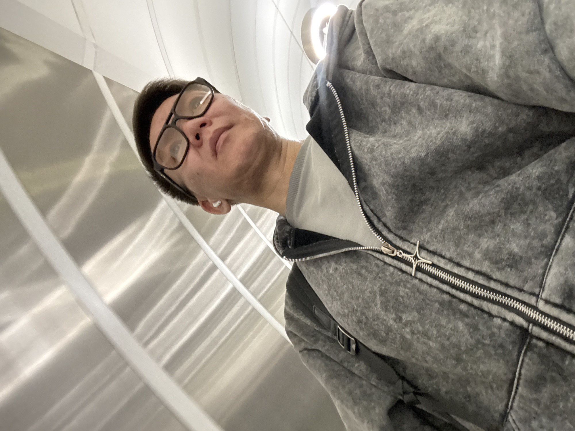 A male individual with short, dark hair and black-framed glasses is captured in a selfie from a low, upward-looking angle, in Moscow, Russia. He wears a light gray t-shirt beneath a dark gray, textured, hooded zipper jacket. A white earbud is visible in his left ear. His expression is neutral as he looks slightly upwards and to the right. The jacket features a silver zipper with a distinctive star-shaped zipper pull. A black strap, likely from a bag, is visible over his right shoulder. The background consists of modern, reflective surfaces, possibly metal panels, displaying vertical lines and bright light reflections, indicative of an indoor, well-lit public space, perhaps a subway station or a contemporary building interior. There is no visible text in the image.