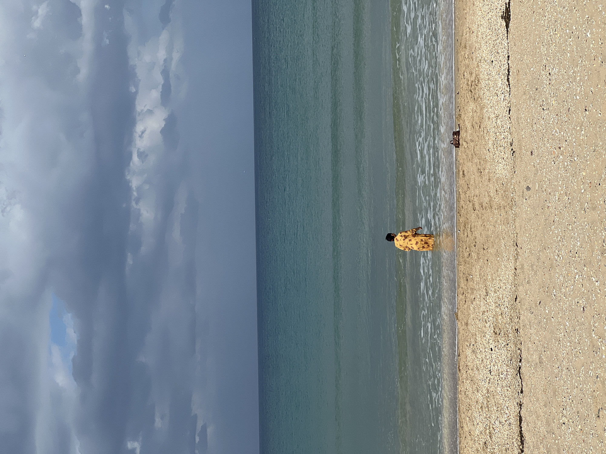 A medium shot depicts a person standing in shallow water at a beach, likely in M'Diq, Morocco, though precise location details are not discernible. The person is wearing a patterned yellow garment and appears to be looking down. The ocean water is a subdued teal color with gentle waves. A sandy beach adjoins the water, transitioning to a coarser, shell-strewn section further inland. Overcast skies dominate the background.