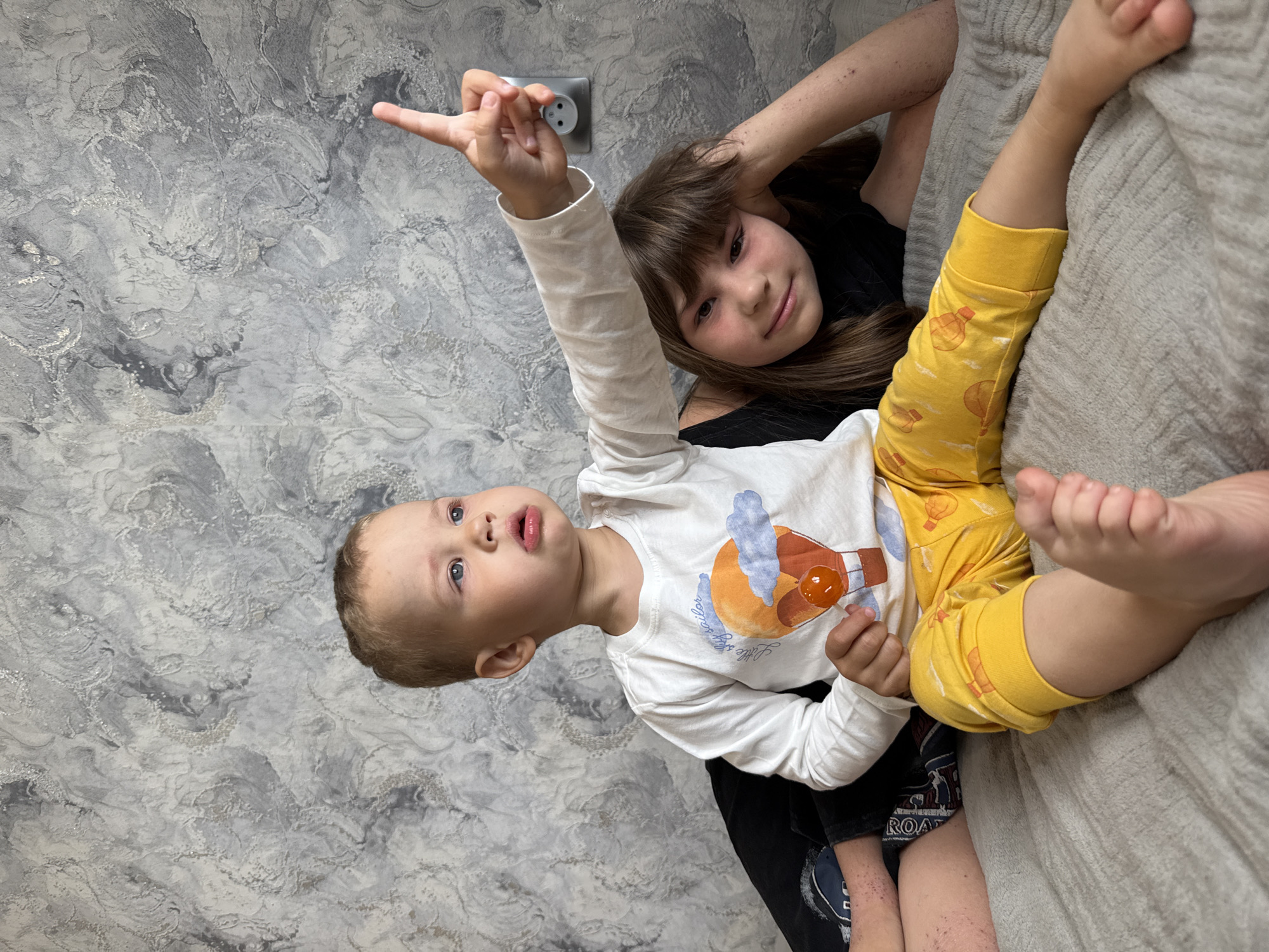 Two children are indoors. A younger child in a white long-sleeved shirt with a hot air balloon design and yellow pants with hot air balloon prints points their index finger upward near an electrical outlet. An older child is behind them, wearing a black shirt. The scene is inside, possibly a home. The background shows a gray and white textured wall. The location is likely Rusanovo, Russia, due to the query's context, but the image itself does not provide proof.