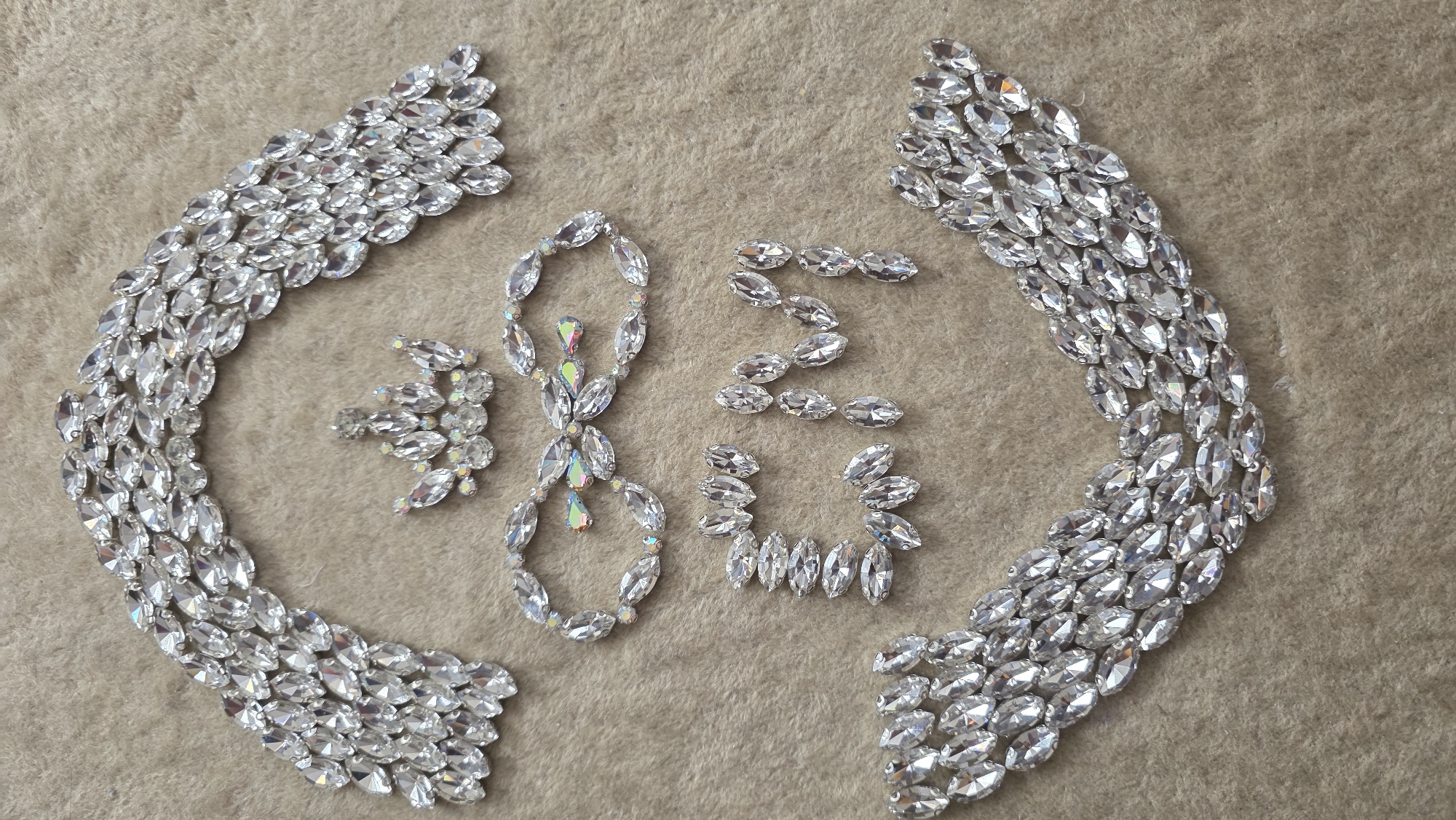 The image showcases an artistic arrangement of numerous sparkling jewels on a light beige, soft-textured fabric surface, possibly velvet or fleece.
The main subjects are clear, marquise-cut rhinestones, with several exhibiting an iridescent (Aurora Borealis) finish, reflecting hues of pink, blue, and yellow. These jewels are meticulously arranged to form various decorative elements.
Two large, dense crescent-shaped formations of jewels are prominent on the left and right sides of the image. Each crescent consists of multiple rows of closely set marquise stones, creating a flowing, shimmering border.
In the central area of the image, jewels are arranged to clearly form the numbers "2" and "0". Around these numbers, there are smaller, intricate clusters of jewels, some with iridescent properties, forming decorative motifs that resemble stylized flowers or ornate embellishments. A particularly detailed cluster with iridescent stones is visible near the center-left.
The activity depicted is the creative arrangement of these jewels, possibly for display, crafting, or as part of a design process. The lighting appears bright and even, highlighting the facets and reflective qualities of the stones. No people are visible, and there are no explicit location cues within the image.