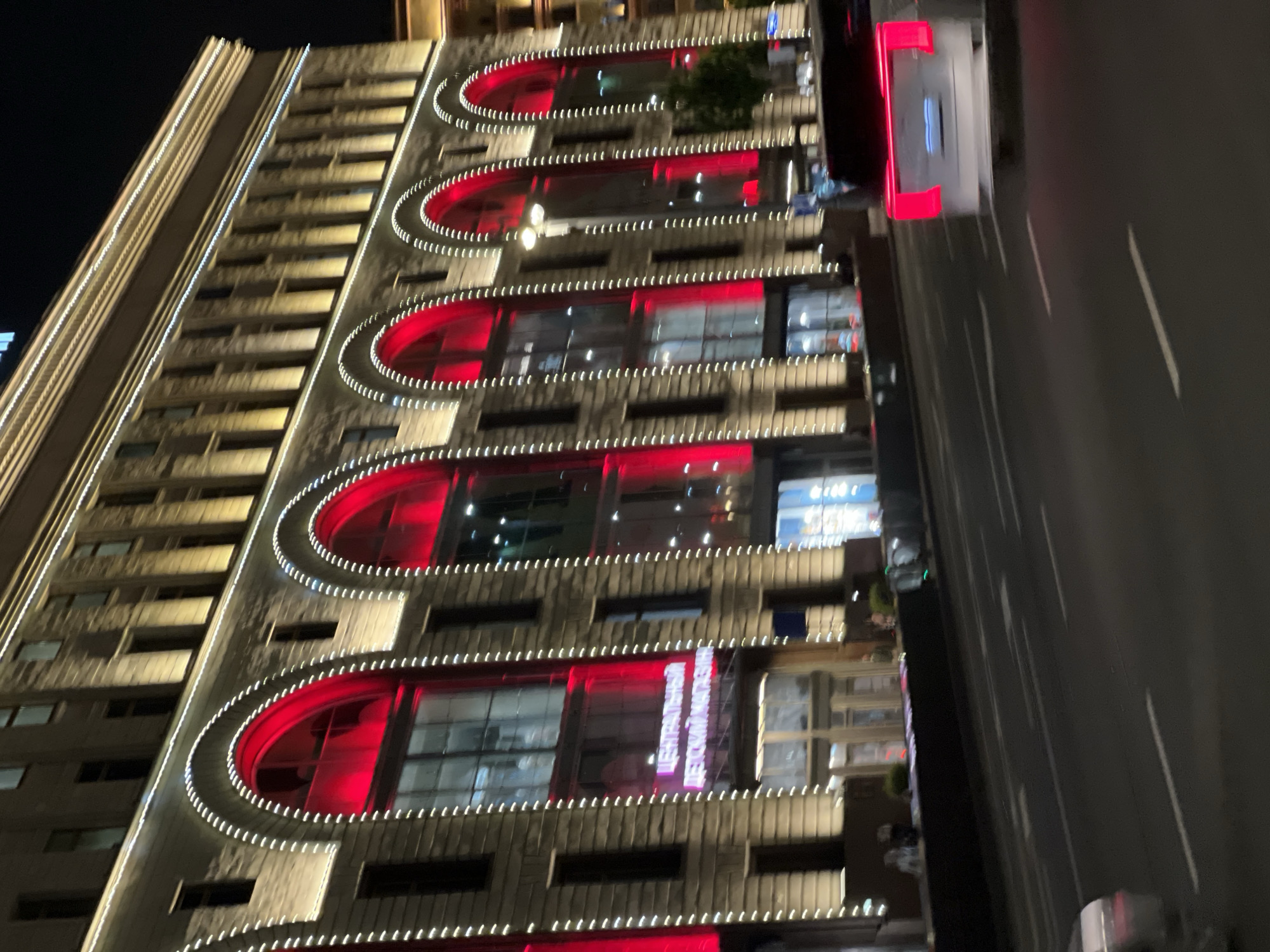 The image depicts the Central Children's Store on Lubyanka Square in Moscow, Russia, at night. The building is illuminated with white string lights outlining its arched windows, which are filled with red light. The upper floors of the building are lit with yellow lights. A street runs in front of the building, with several cars visible. One car in the foreground has its brake lights illuminated in red. The scene is slightly blurred due to motion, suggesting the photograph was taken from a moving vehicle.