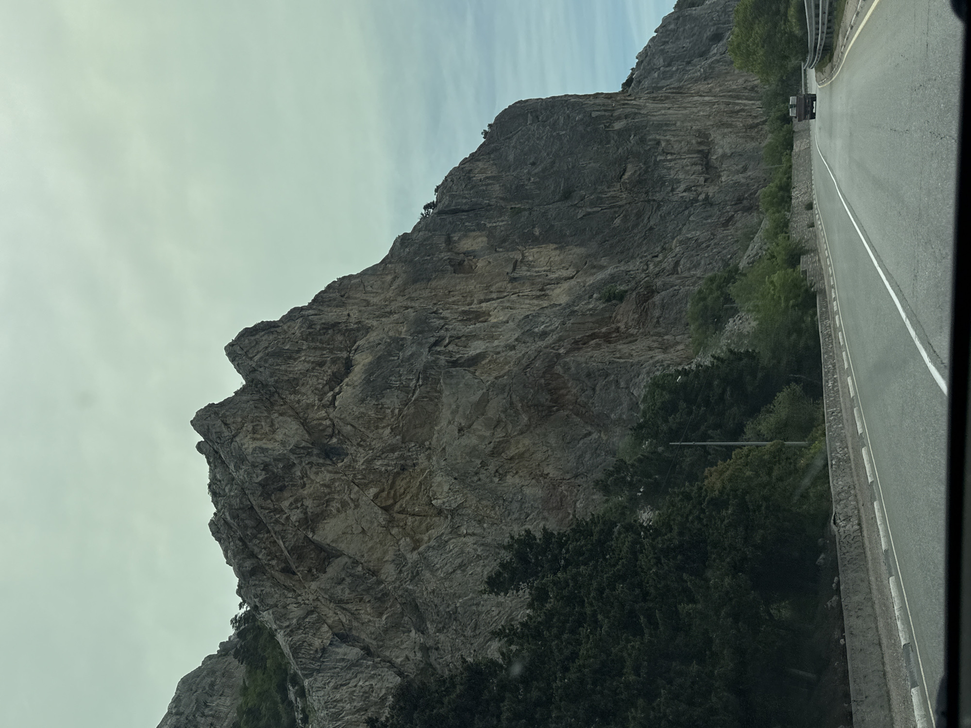 The image shows a large, rocky mountain face next to a road. The road has a white line dividing the lanes and a concrete barrier along the side closest to the mountain. Trees and vegetation grow at the base of the mountain and along the roadside. A vehicle is visible further down the road. The sky is overcast. The location is Орлиновский округ, Ukraine.