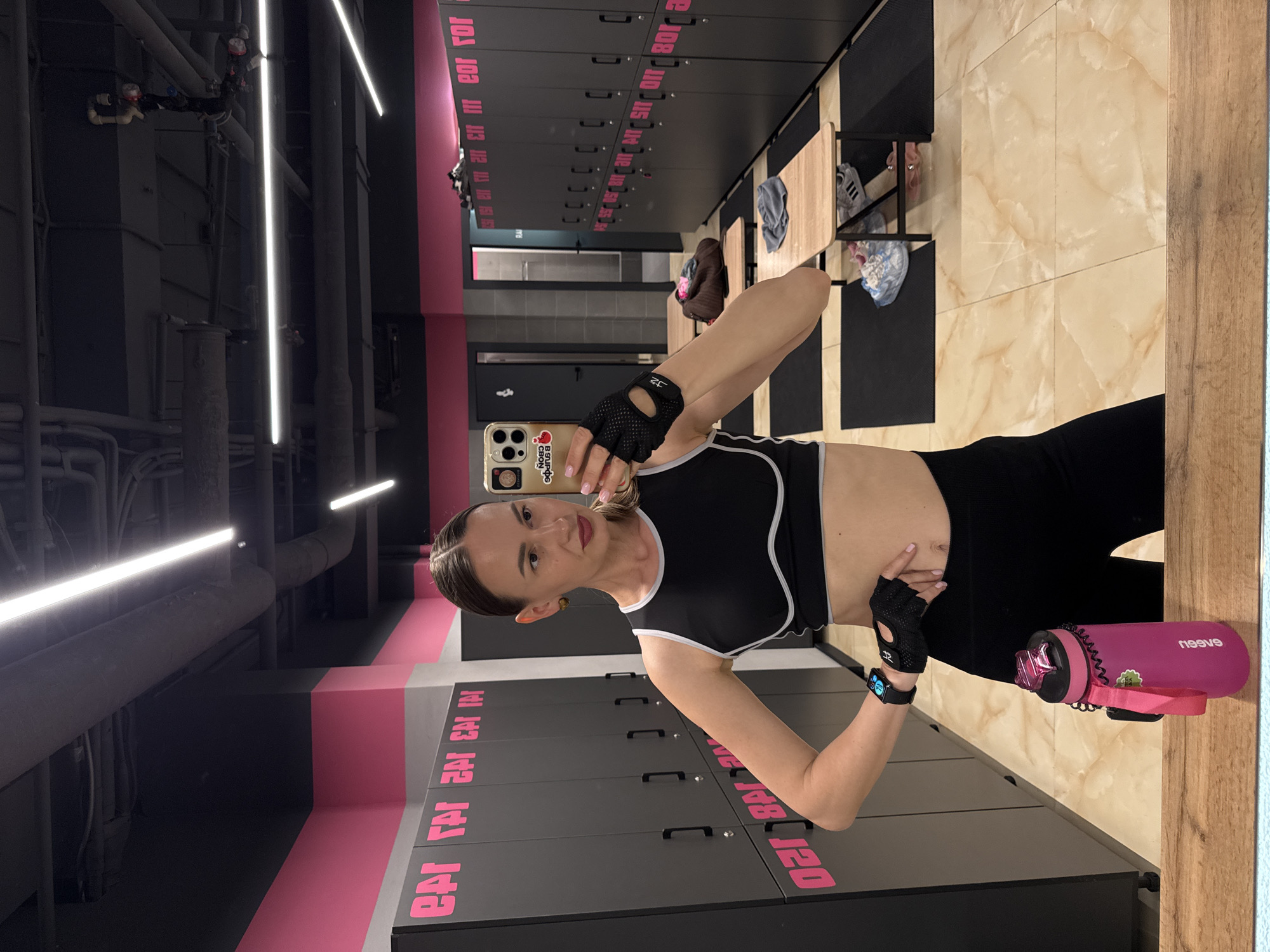 An indoor, vertically oriented mirror selfie captures a woman posing in what appears to be a modern gym changing room or locker facility. The woman, holding a gold-colored iPhone with a cherry-themed pop socket, is seen from the chest up, with her left hand placed on her midsection and her right arm extended to hold the phone. She is wearing a black sports bra with white trim, black leggings, and fingerless gym gloves, along with a smartwatch on her left wrist. Her hair is pulled back, and she looks directly at the camera.
The background reveals rows of dark grey lockers, some adorned with bright pink numbers and stylized text. Visible reversed text on the lockers includes "11 114 115 110 108," "140 141 142 143 141," and "120-148," indicating locker numbers. Other reversed pink text like "eor for," "en im," "selen," and the Cyrillic word "эфала" is also present, suggesting the location context of Sochi, Russia. A vibrant pink stripe with text that seems to read "EAT EAT EAT IN" runs along a wall. The floor features light brown and beige marbled tiles. Exposed dark pipes and sleek white linear LED lights are visible overhead, contributing to an industrial yet contemporary aesthetic. To the woman's right, a pink water bottle with the reversed brand name "neeguv" (likely Vogueen) rests on a surface. Wooden benches and personal items like clothes and shoes are scattered in the background. The scene is brightly lit by artificial light, and the overall mood is active and functional, typical of a fitness environment.