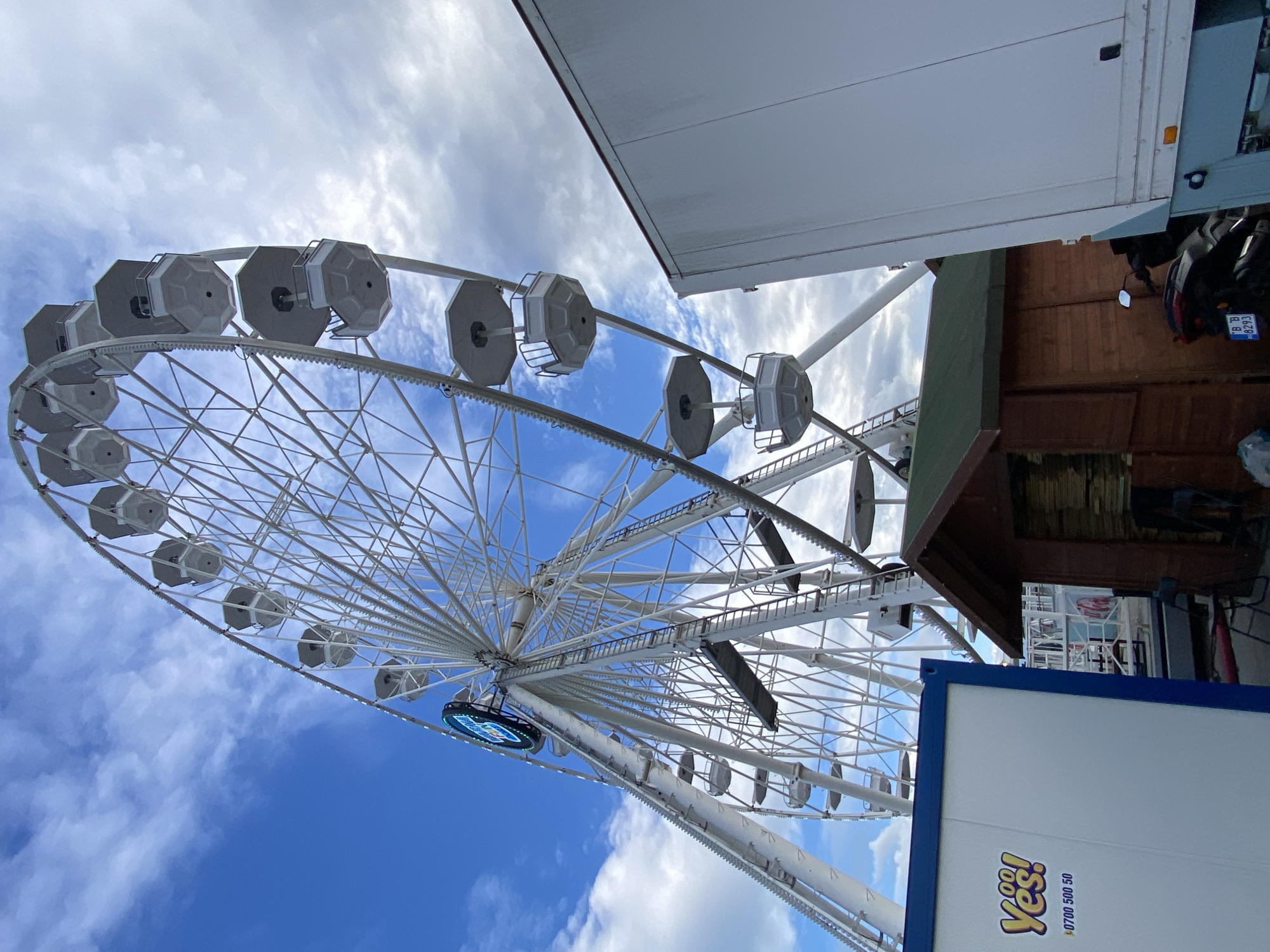 The image depicts a Ferris wheel against a blue sky with scattered clouds. The Ferris wheel is white with enclosed passenger cars. Sections of buildings and vehicles frame the right side of the image. The word "Yes!" is visible in the lower right corner of the image, on what appears to be the side of a truck. The registration plate of a motorcycle parked next to a wooden structure is partially visible in the background. Based on the location information, the image depicts a Ferris wheel in Varna, Bulgaria.