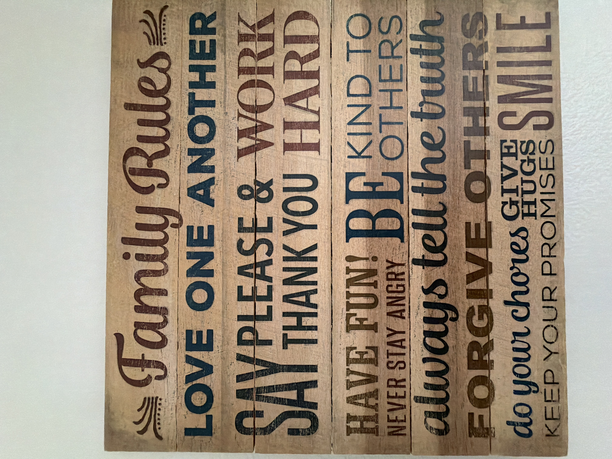 A wooden sign with family rules is mounted on a white wall. The sign is made of horizontal wooden planks. The rules are printed in various fonts and colors, including dark blue, dark red, and black. The rules read: "Family Rules," "LOVE ONE ANOTHER," "SAY PLEASE & WORK HARD," "SAY THANK YOU," "HAVE FUN! BE KIND TO OTHERS," "NEVER STAY ANGRY," "always tell the truth," "FORGIVE OTHERS," "do your chores GIVE HUGS SMILE," and "KEEP YOUR PROMISES." The sign is located in Wellford, United States of America.