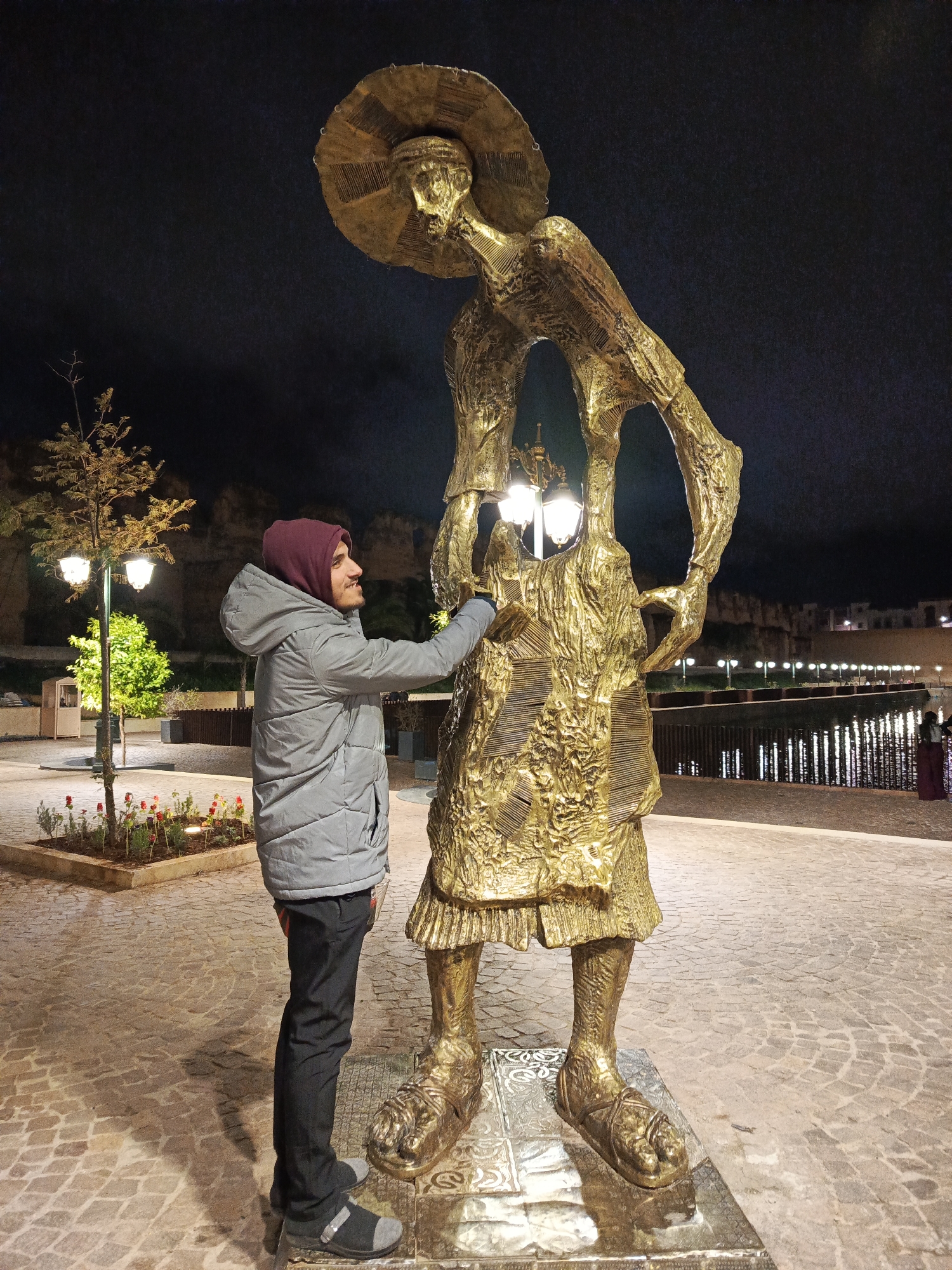 The image captures a man standing next to a large golden sculpture in a public space at night. The man is wearing a grey hooded jacket and dark trousers. He is looking at the sculpture and has his hand extended towards it.
The sculpture is of a stylized human figure with an oversized hat and appears to be crafted from textured metal. The setting appears to be a paved plaza with trees, flowers, and streetlights. In the background, there is a body of water with a railing and lights along the shore. Buildings are visible in the distance. The location is Al Machouar – Stinia, Morocco.