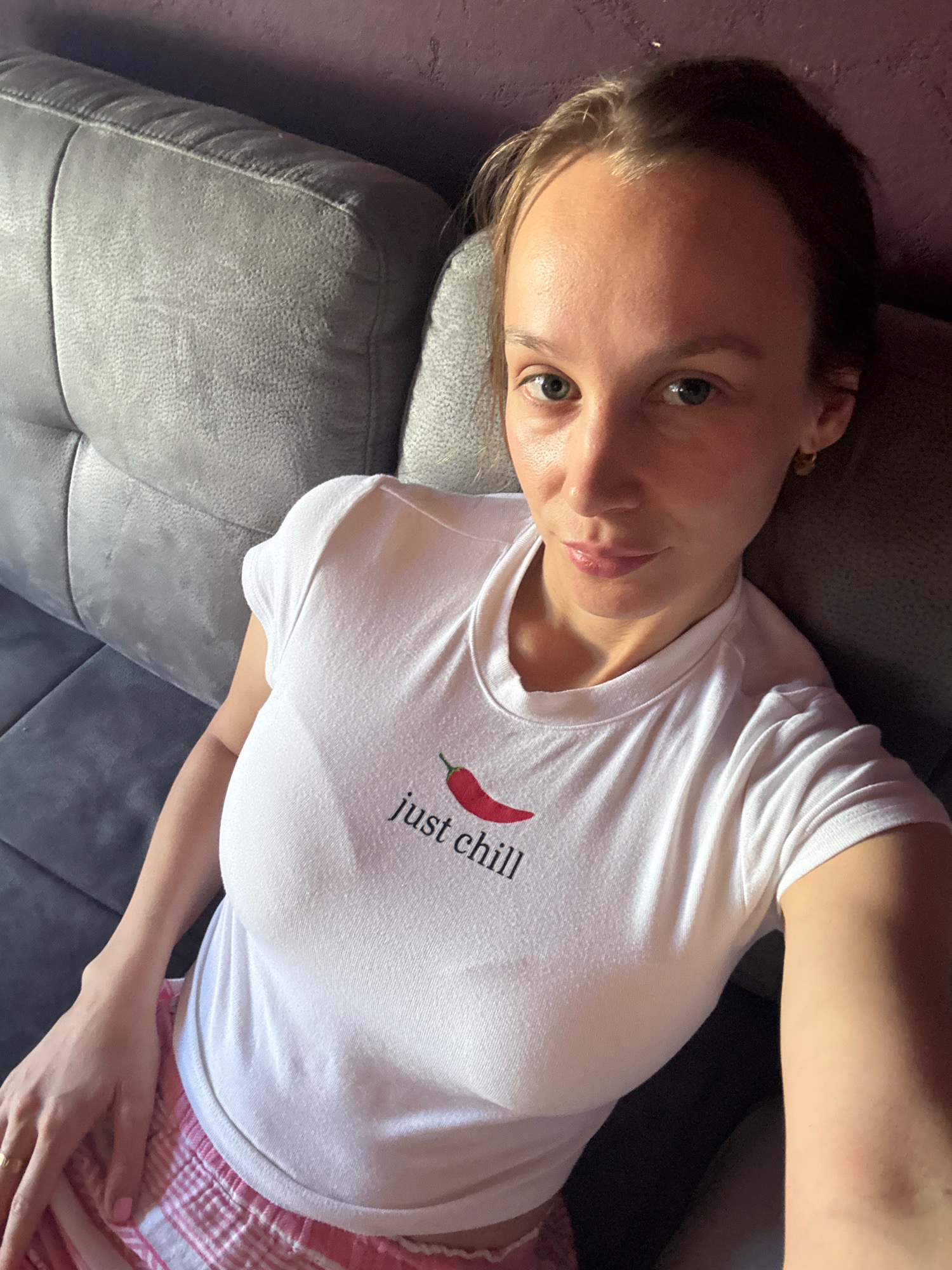 The image shows a woman with light brown hair and blue eyes. She is wearing a white t-shirt with a red chili pepper graphic and the words "just chill" printed on it, and pink plaid shorts. She is sitting on a gray tufted sofa in what appears to be a living room. The wall behind her is a dark purple color. There is no indication of the location being in Überherrn, Germany.