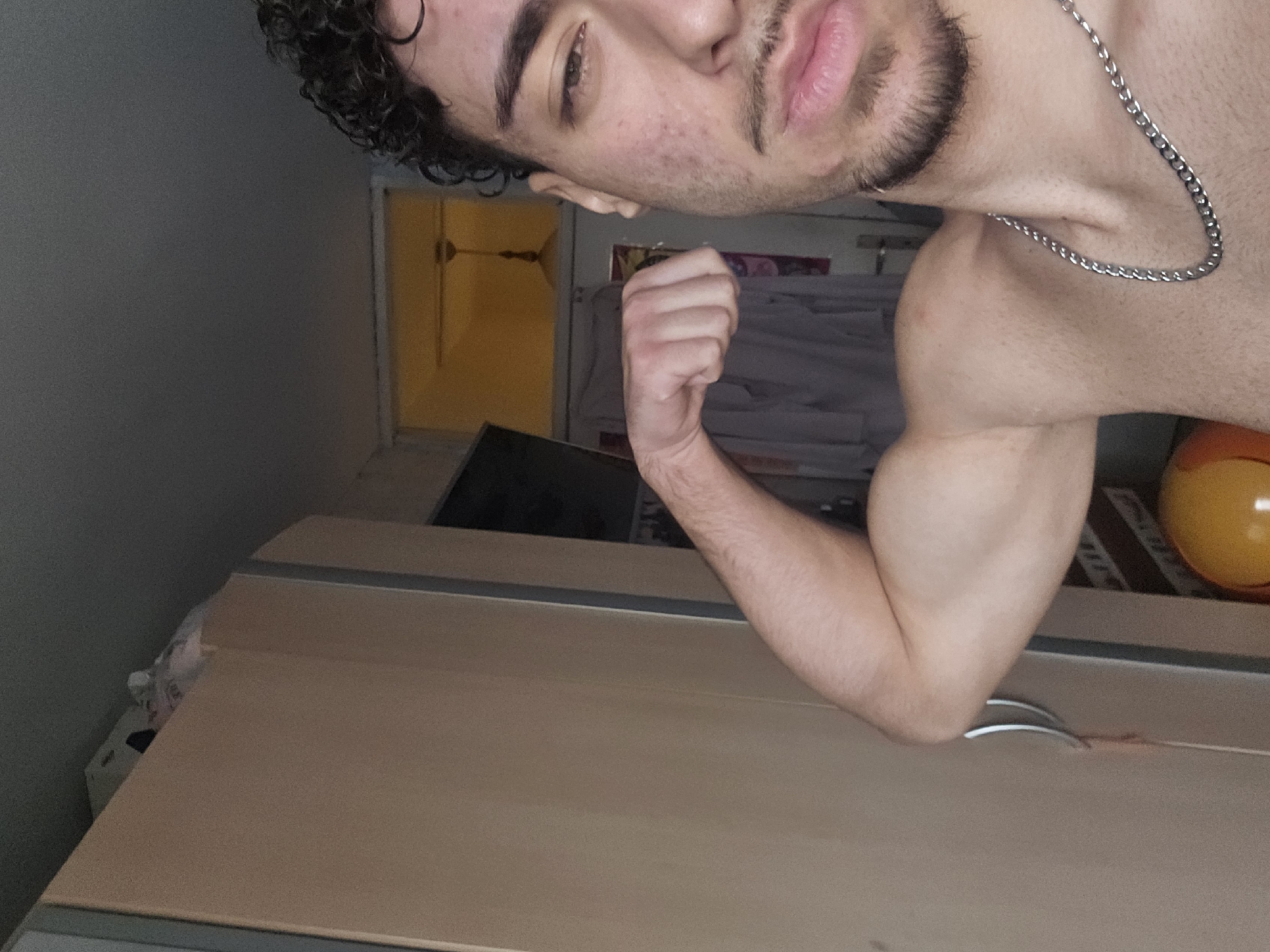 The image displays a shirtless male subject, appearing to be a young adult, captured from the chest up in an indoor setting. The subject has light skin, dark curly hair visible on the right side of his head, stubble on his chin and upper lip, and a silver chain around his neck. He is actively flexing his left biceps, with his left arm bent at the elbow and his fist clenched. The subject's face, visible on the right half of the frame, shows visible blemishes or acne on his cheek. His right eye is partially closed, and his gaze is directed towards the viewer.
The background on the left features a light brown cabinet or wardrobe with a silver handle. A white box or object is perched on top of this cabinet. To the right of the subject's arm, an open doorway leads to an adjacent room, illuminated by a warm, yellowish light emanating from a ceiling fixture, possibly a fan or chandelier. Some light-colored garments appear to be hanging on a hook or doorframe within this adjacent space. A partially visible orange and yellow spherical object is present in the lower right foreground. There are no discernible elements within the image to identify a specific location within the city of Sofia, Bulgaria.