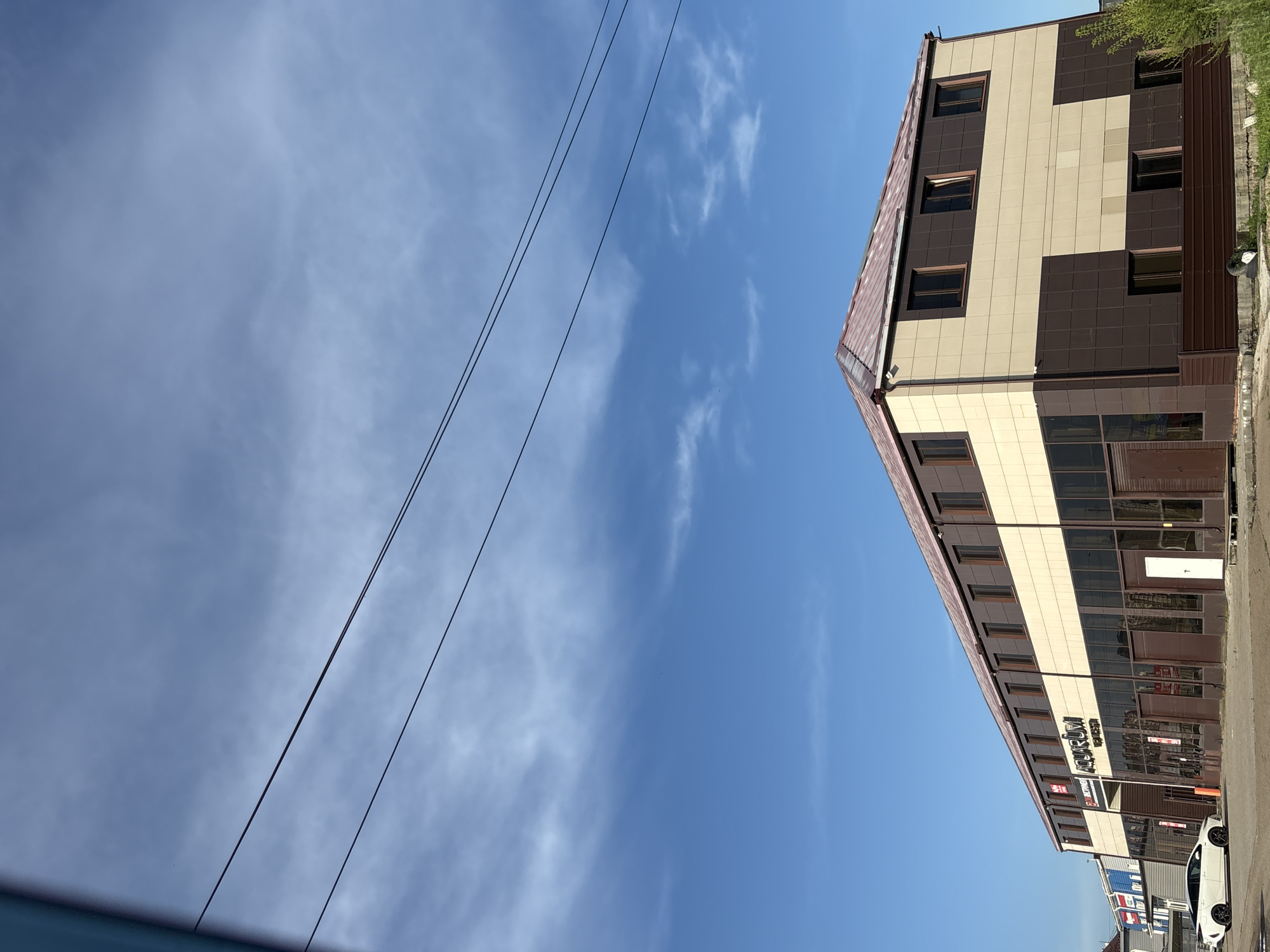 The image shows a two-story commercial building with a light beige and dark brown tiled facade, under a clear blue sky with scattered white clouds. The building has a reddish-brown sloped roof and numerous windows. A white sedan is parked in front of the building, on a paved surface. The building has signage that appears to read "AQUARIUM". The location is identified as Blagoveshchensk, Russia. The scene appears to be an ordinary street view of a commercial establishment.