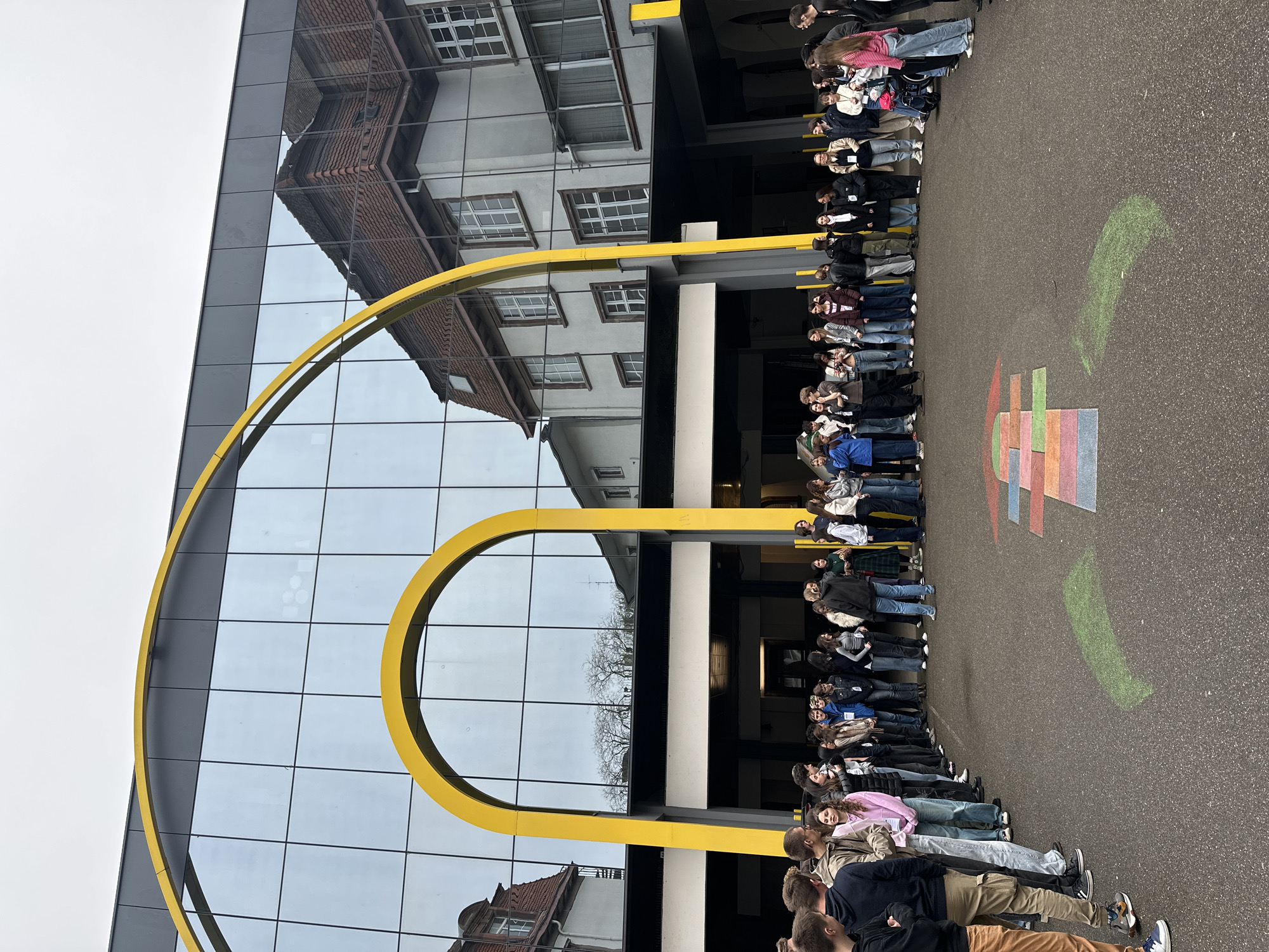A long row of people stand outside a modern building with a large yellow arch in front. The building is mainly glass, reflecting a traditional building and the sky. A colorful rectangular design is drawn on the pavement. The scene is likely in Strasbourg, France, given the architectural style of the reflected building.