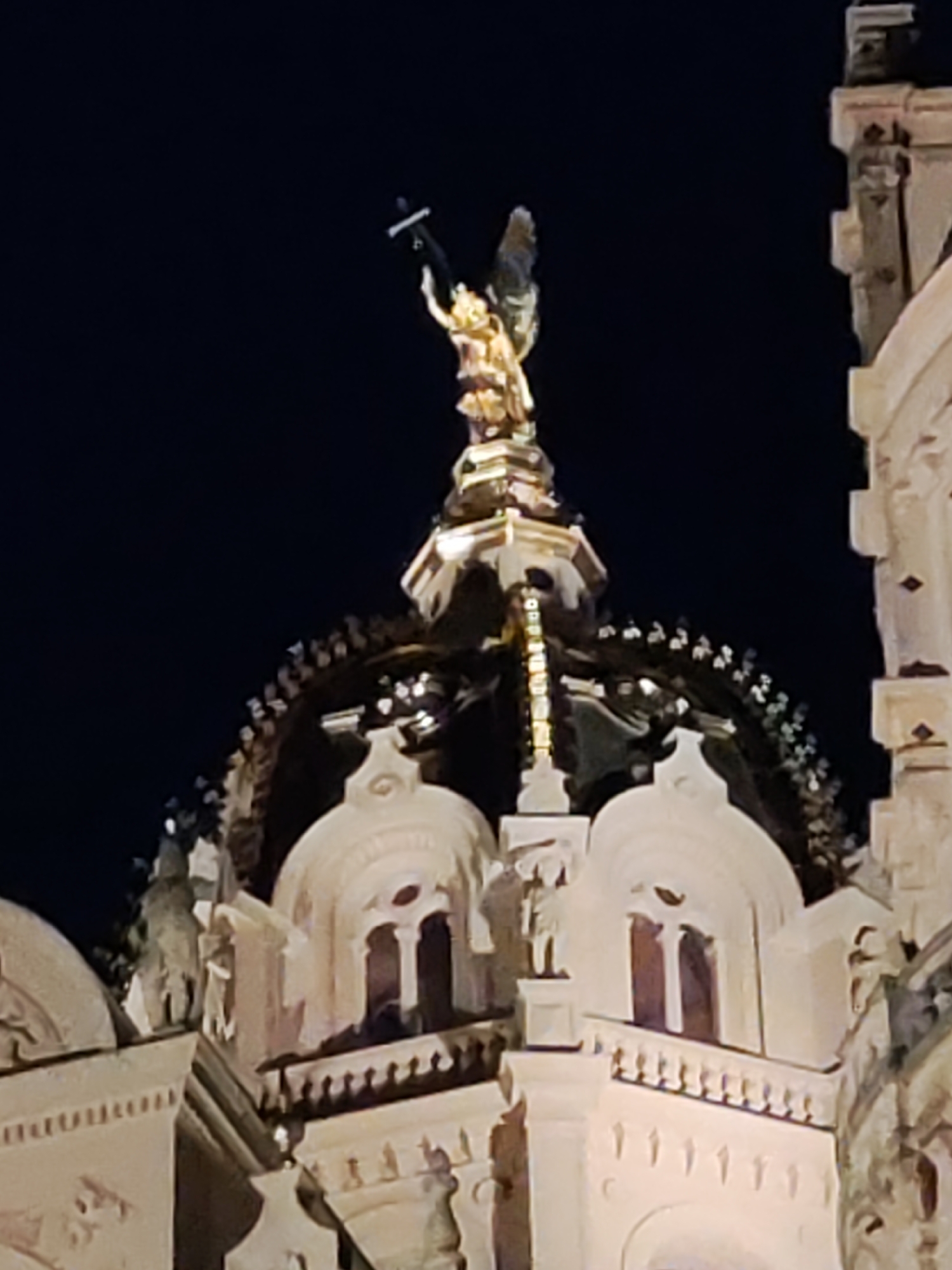 The image shows the gilded statue of an angel at the top of the dome of Schwerin Cathedral in Schwerin, Germany. The angel is holding a cross in its right hand and is depicted with wings. The statue is illuminated against a dark night sky. The architecture of the cathedral, with its ornate details and arched windows, is visible below the statue. The image captures the angel from a low-angle perspective.