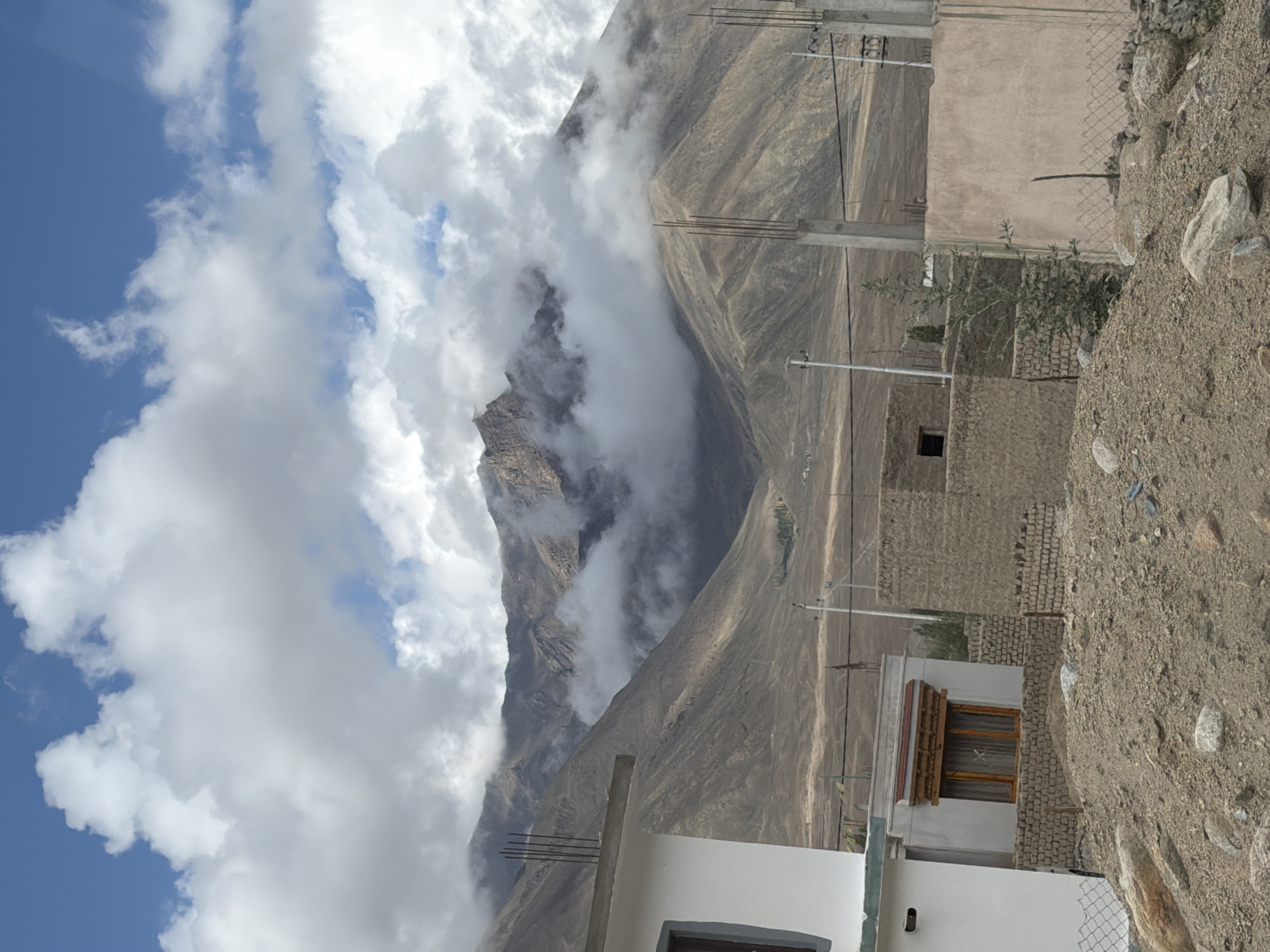 The image captures a scene in Spituk, India, characterized by a dramatic mountainous landscape partially obscured by clouds. In the foreground, the rocky, uneven terrain is dotted with boulders and dirt. Several structures are visible: a white building with a grey trim and visible rebar protruding from its roofline occupies the left side of the frame. Adjacent to it is a smaller, multi-story building constructed with brick and plaster, featuring a wooden-framed window with curtains. Further to the right, another building constructed from rough-hewn bricks is present. Utility poles with wires are scattered throughout the mid-ground, and a chain-link fence runs along the lower edge of some of the properties. The background is dominated by imposing, barren mountains, their slopes etched with shadows and light, with dramatic cloud formations swirling around their peaks. The sky above is a bright blue, interspersed with fluffy white clouds.