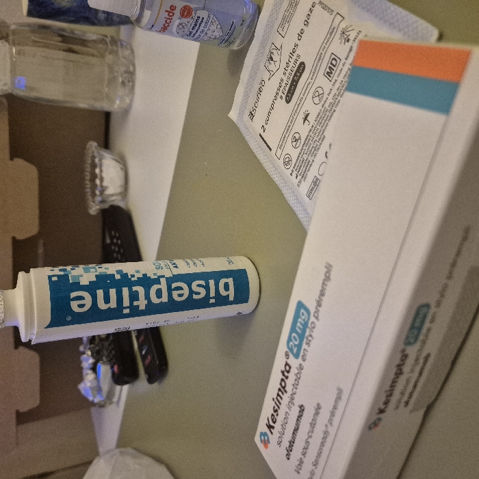 The image displays several medical items on a table. There's a bottle of "biseptine," an antiseptic solution, alongside a box of "Kesimpta 20 mg," an injectable solution pre-filled in a pen, containing ofatumumab for subcutaneous use. A small bottle labeled "baccide" is visible, identified as hand sanitizer. Additionally, the image shows a sterile gauze pad with the markings "2 compresses stériles de gaze," indicating two sterile gauze pads with a size of 7.5 x 7.5 cm.