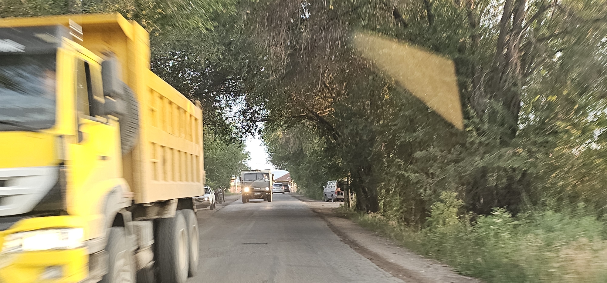 The image depicts a road scene in the city of Жамбыл, Kazakhstan, characterized by a canopy of trees overhanging the asphalt surface. In the foreground, a large, yellow dump truck is captured in motion, with its front section prominently visible on the left side of the frame, indicating the perspective is from within another vehicle. The truck's tires and the lower part of its chassis are visible, blurred due to movement. Further down the road, another truck, this one military green and appears to be a KAMAZ model, is traveling towards the viewer. Several other vehicles, including a white car and a smaller silver vehicle, are also present on the road. The road itself appears to be a two-lane asphalt surface with a dirt shoulder on the right, lined with dense green trees and vegetation. A distinctive rectangular yellow reflection is visible on the right side of the frame, potentially from a surface within the camera's field of view. The lighting suggests it is daytime, possibly late afternoon or early evening, with sunlight filtering through the tree canopy.