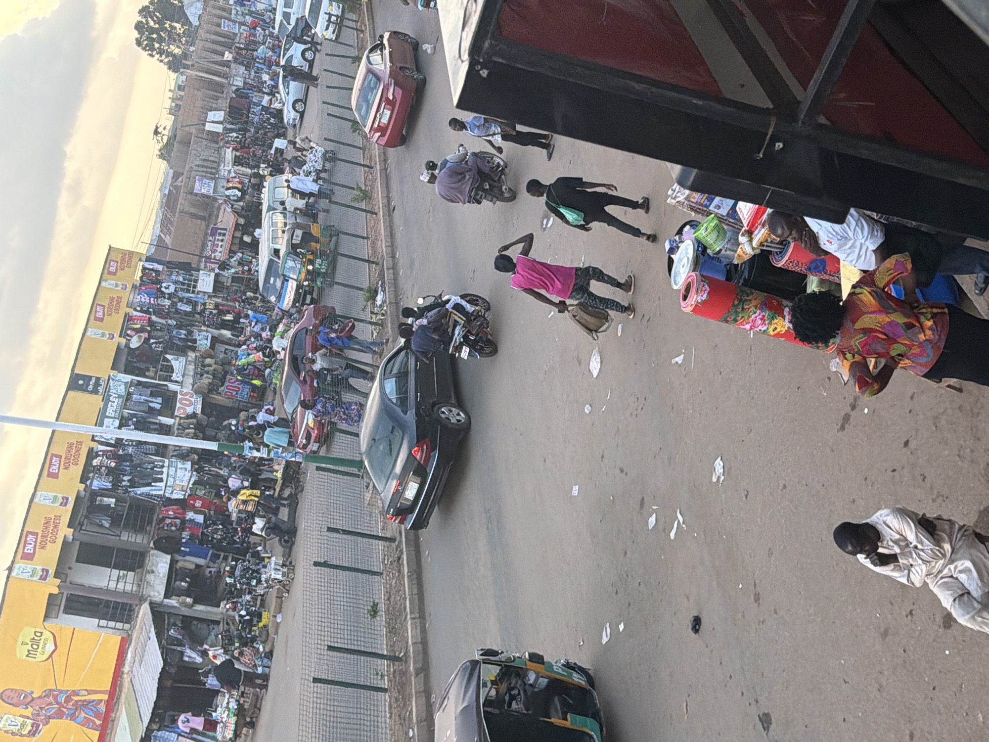 This media file captures a bustling street scene in Bukuru, Nigeria, likely during the late afternoon or early evening, as suggested by the warm, diffused lighting and soft shadows. The image is a top-down perspective, looking onto a busy road lined with market stalls and shops.
The foreground is dominated by a section of a market with various goods displayed, including rolled carpets and what appear to be textiles. Several people are visible, some interacting with the goods and others moving through the market. One person in a light-colored garment is kneeling, possibly in prayer or resting.
Further into the scene, the road is filled with activity. Vehicles, including cars, motorcycles, and a three-wheeled auto-rickshaw, navigate the crowded street. People are walking and gathering around the shops and stalls. The shops themselves appear to be selling clothing and other merchandise, with signs featuring text like "ENJOY NOURISHING GOODNESS" and "Malta GUINNESS." Some of the shops have yellow signage with red lettering.
The overall impression is one of a vibrant and active marketplace, characteristic of a busy urban environment in Nigeria. The atmosphere appears dynamic and lively, with the everyday commerce and movement of people. The sky visible in the upper left corner is a pale, hazy yellow, indicating the time of day and possibly a warm, humid climate. The image is oriented vertically, with the top of the scene appearing on the left.