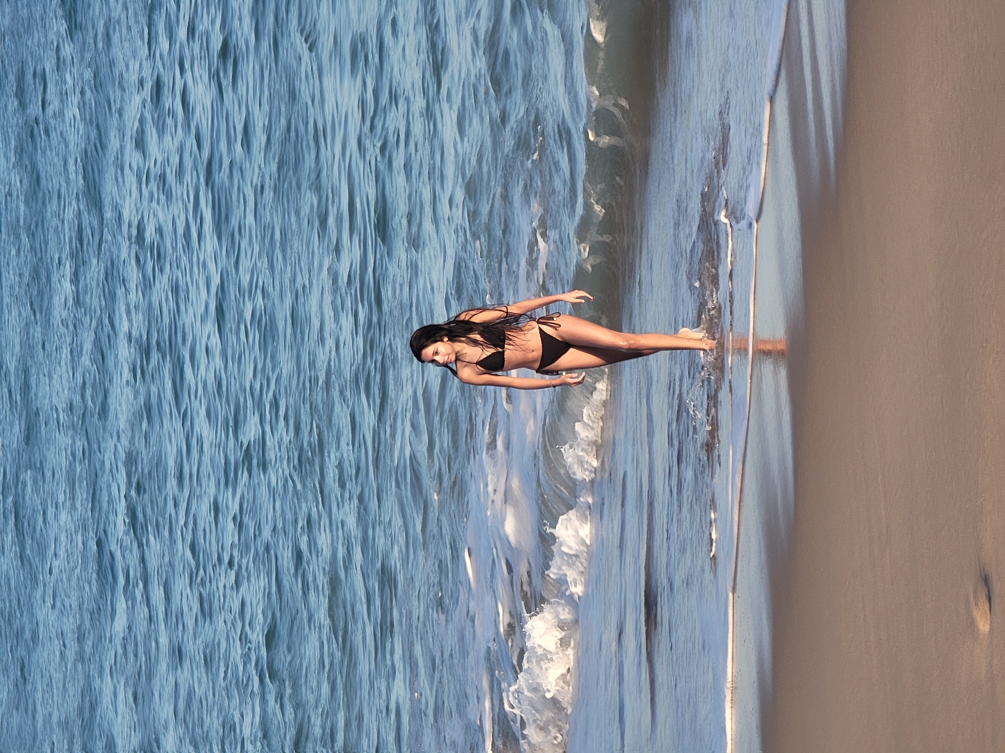 A young woman in a black bikini walks through the shallow ocean water near the shore. The woman has dark hair and is facing to her left, looking out at the water. The water is blue and wavy, with white foam from the waves washing onto the sand. The sand is tan and wet at the water's edge. The scene is likely on a beach in Quarteira, Portugal, given the typical appearance of the coastline and water.