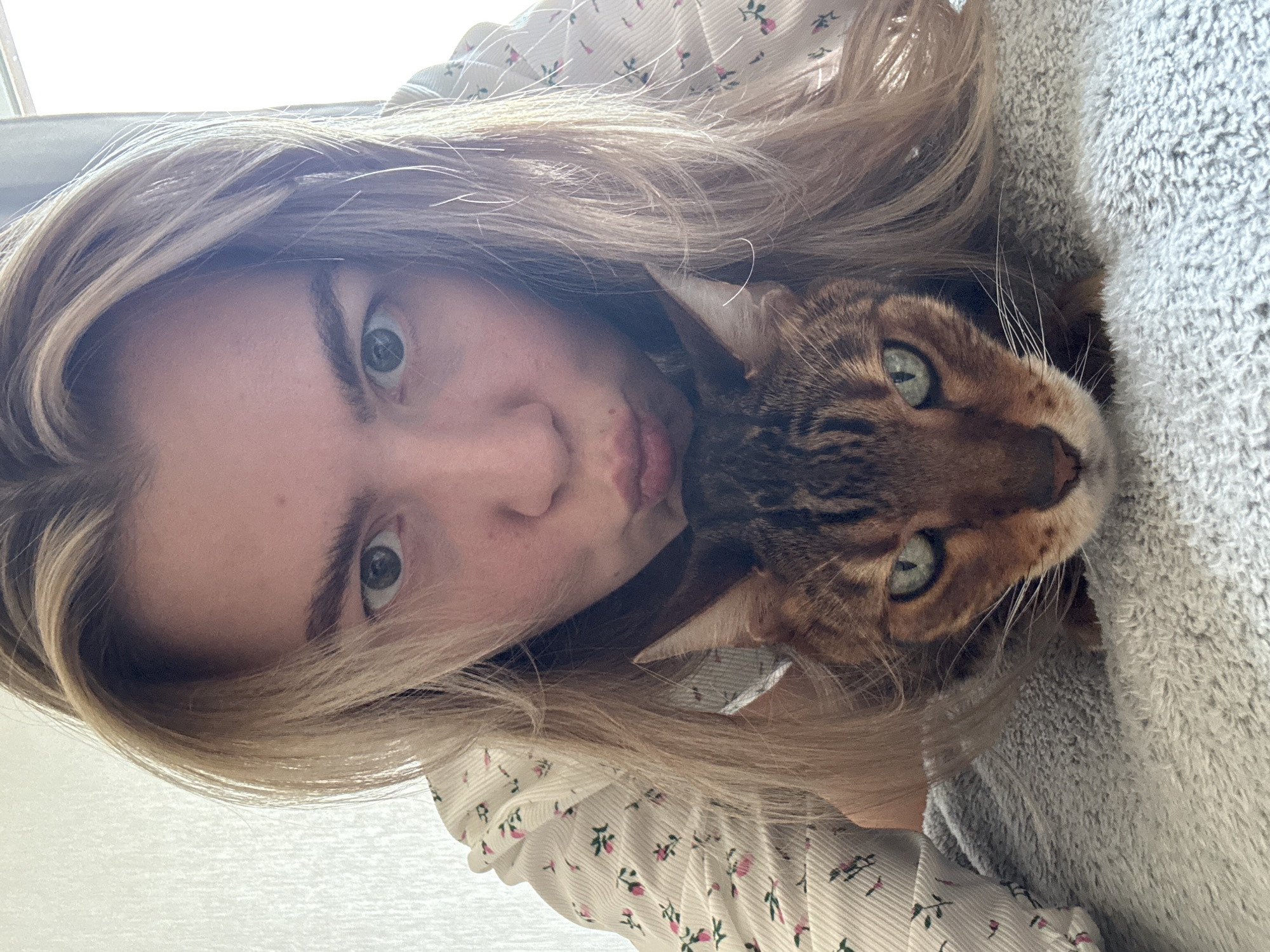 A close-up portrait, oriented sideways, captures a person and a cat in an intimate and affectionate pose, likely a selfie. The primary subject on the left is a young woman with long, flowing light brown or blonde hair that cascades around her face. She has wide, light-colored eyes (appearing green or blue-green) and a playful, slightly puckered or "kissing" expression directed towards the cat. She is wearing a light-colored top adorned with a small, repeating pink floral pattern.
Cuddled closely beside her, and partially nestled within her hair, is a domestic cat with distinctive spotted and striped (marbled) fur, characteristic of a Bengal breed. The cat also has striking green eyes and prominent whiskers, and its gaze is directed towards the camera.
The scene appears to be set indoors during daytime, indicated by the bright, somewhat overexposed lighting in the background, suggesting a window or strong light source. On the right side of the frame, a textured, light grey fabric, possibly a blanket or soft furnishing, is visible, enveloping part of the cat. The overall impression is one of companionship and warmth between the two subjects. There is no visible text in the image.