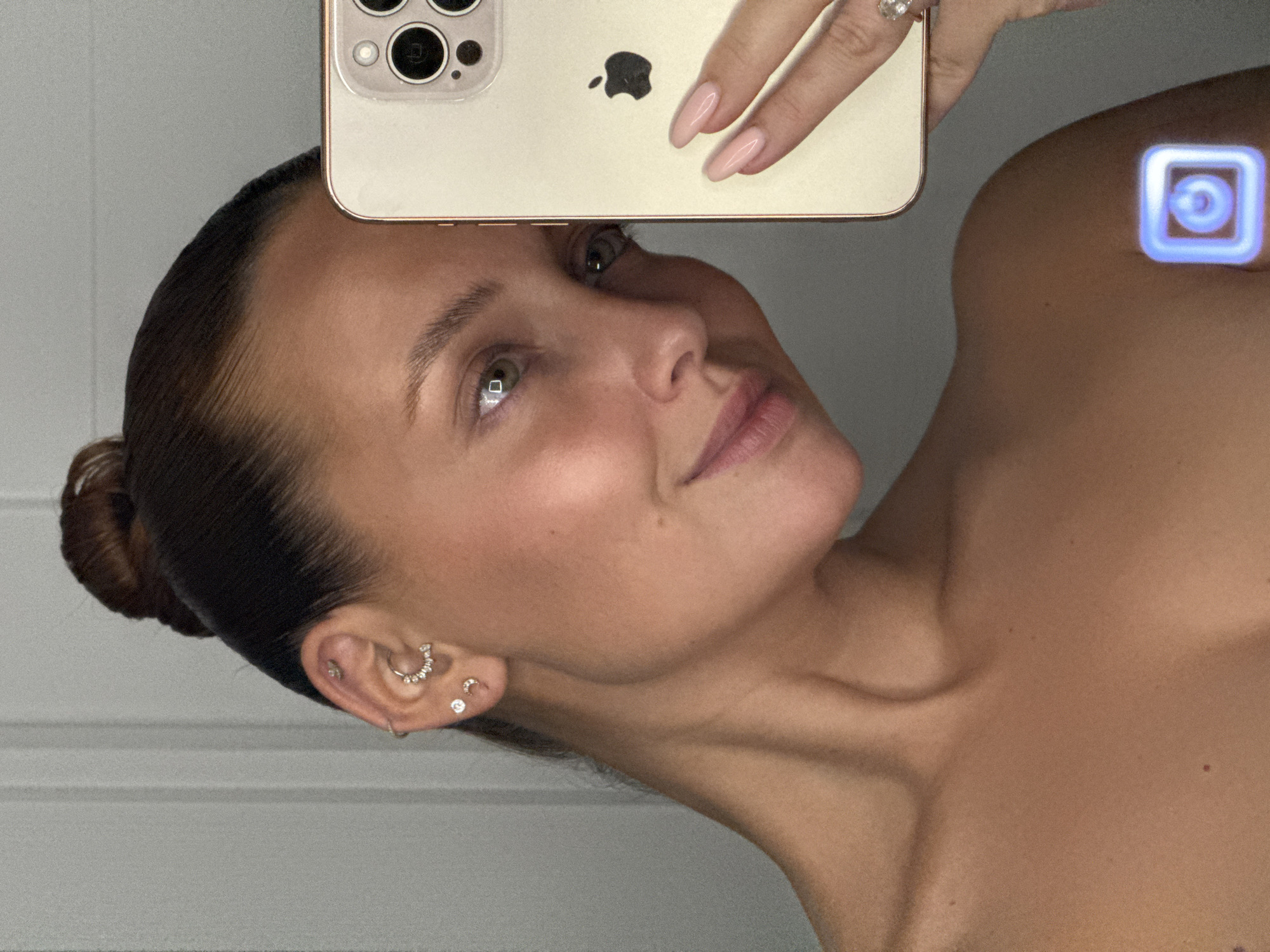 A woman is taking a selfie in what appears to be a bathroom or dressing area. She is holding a gold iPhone up to take the picture, and the camera is angled to capture her face and shoulder. She has dark hair pulled back in a bun and is wearing multiple earrings in her left ear, including a hoop and studs with small diamonds. Her nails are painted a pale pink, and she is wearing a diamond ring on her left hand. There is a blue light projection on her shoulder. The background is a plain, light-colored wall, possibly a mirror or door. The image is a close-up, focusing on the woman's face and upper body. There is no information in the image that allows for the identification of a specific location in Chaikovsky, Russia.