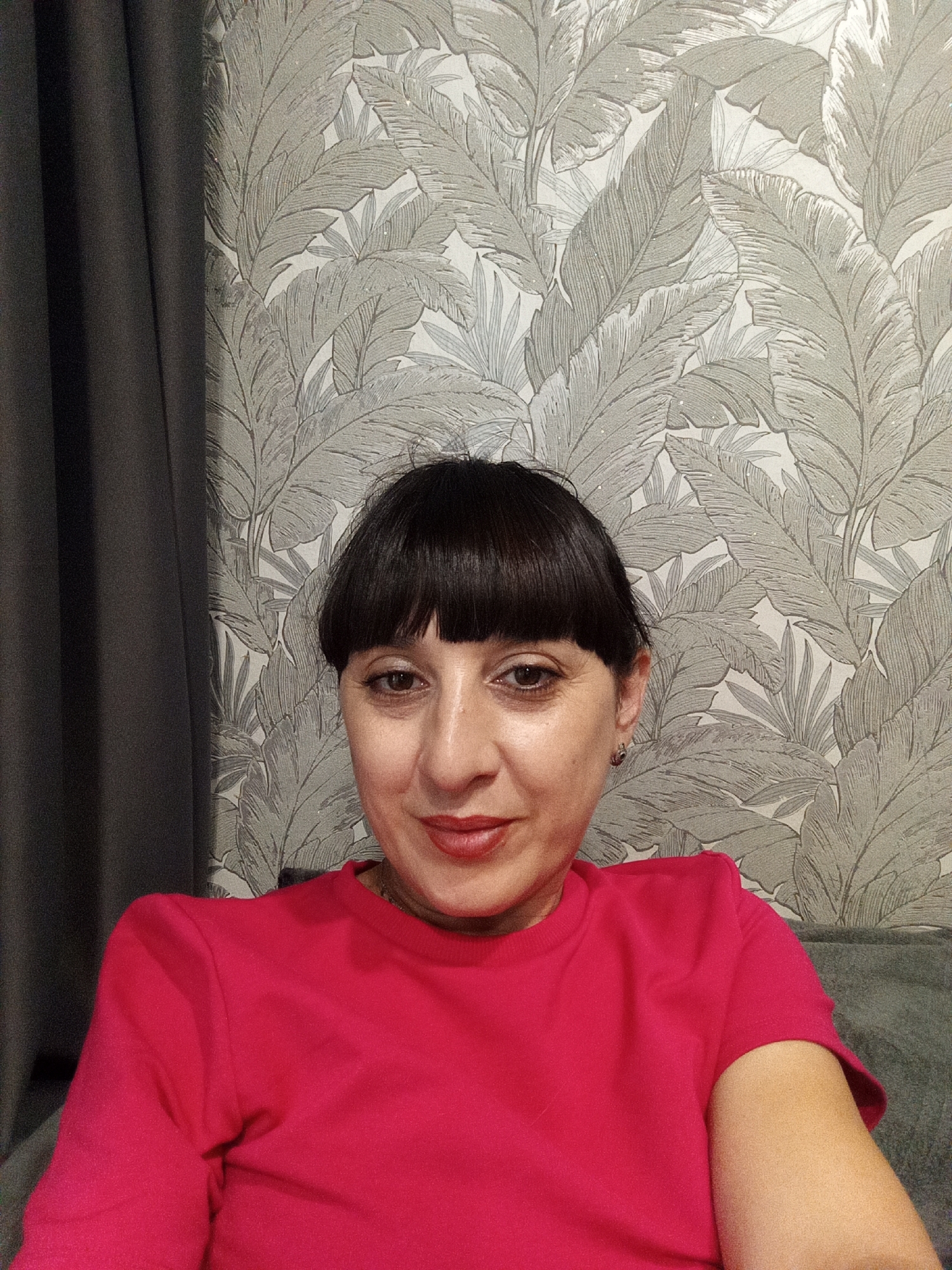 A medium shot portrait of a woman is captured indoors. She is positioned centrally in the frame, facing the camera directly. The woman has dark, straight hair with bangs cut straight across her forehead, and her dark eyes gaze forward. She is wearing a bright pink crew-neck shirt. Her makeup includes dark eyeliner and red lipstick. To her left, a dark gray curtain hangs, partially obscuring the background. The background behind her head and to her right features wallpaper with a pattern of large, light gray tropical leaves on a white base. The lighting appears to be artificial and originates from the front, casting minimal shadows. The overall setting suggests a home interior, likely a living room or bedroom. No specific landmarks or elements identifying the city of Belogorsk, Russia, are visible in the image.
