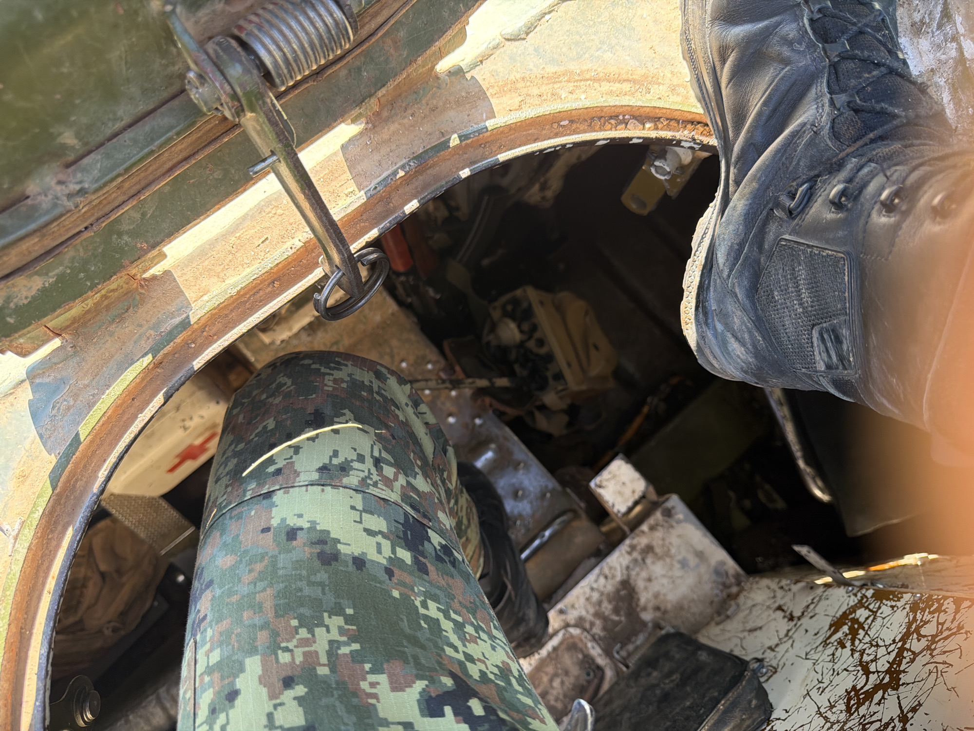 This close-up, high-angle shot captures a person inside a military vehicle, likely a tank, based on the interior components visible. The subject is wearing camouflage pants and black combat boots, suggesting military personnel. Their legs are bent, indicating they are either seated or preparing to move within the confined space.
The foreground is dominated by the subject's legs and boots. The camouflage pattern on the pants features shades of green, brown, and black. The black boots appear worn and sturdy. To the left, a metallic hatch mechanism with a spring is visible, suggesting an access point to the vehicle's interior. The interior itself is dimly lit and cluttered with various mechanical components, wires, and what appears to be fabric or equipment bags. A red cross symbol is faintly visible on a white surface in the background, possibly indicating medical supplies or a first-aid station within the vehicle.
The scene is industrial and utilitarian, consistent with a military vehicle. The lighting suggests it could be daytime, with natural light entering from the open hatch. The presence of dust and wear on the interior surfaces indicates the vehicle is operational and has likely been used in the field. The overall impression is one of a functional, operational military environment. The location context of Beliakovets, Bulgaria, places this scene within a specific geographical and military setting. No discernible text is visible in the image.
