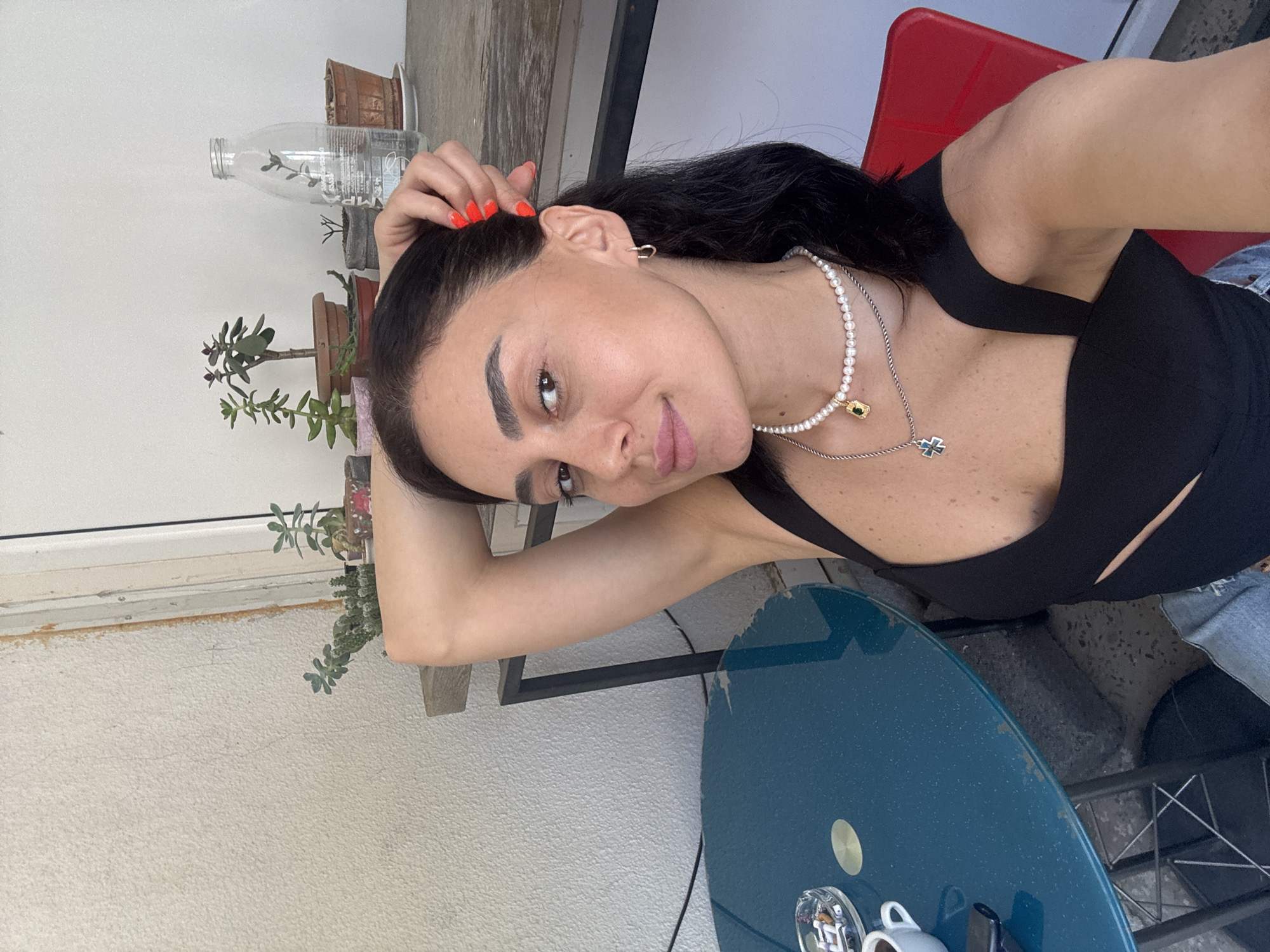 A woman with dark hair is taking a selfie in a bright, outdoor setting, likely a balcony or patio. She is wearing a black crop top, jeans, and two necklaces, one a pearl choker and the other a silver chain with a cross pendant. Her right arm is raised, with her hand resting on her head, and her left arm is bent with her hand near her chest. Her fingernails are painted bright red.
In the background, there is a blue circular table with a coffee cup, an ashtray with several cigarettes, and what appears to be a vape pen. Behind the table, a black metal chair is visible, and further back, a shelf holds several potted plants and a glass bottle of water. The wall behind the plants is textured and light-colored. The scene suggests a casual, relaxed atmosphere. While the specific location in Tbilisi, Georgia, cannot be determined from the image alone, the presence of outdoor seating, potted plants, and casual attire are common elements found in cafes, homes, or guesthouses throughout the city, particularly in areas with outdoor living spaces.