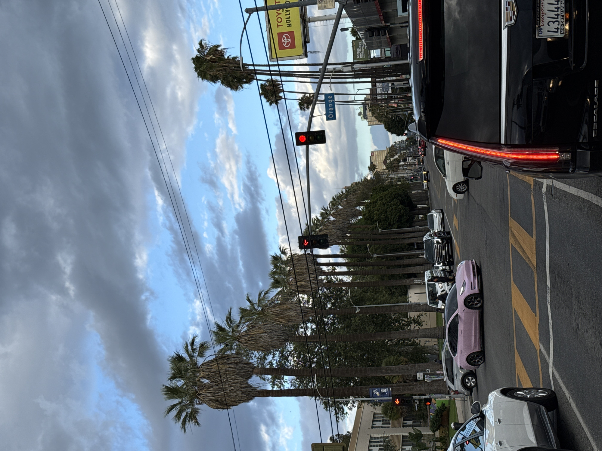 Captured in West Hollywood, United States, the image portrays a city street scene. Palm trees line the avenue under a sky filled with clouds. Traffic is heavy, with a variety of cars visible, including a pink Tesla, a black Cadillac Escalade with license plate '43444', and a white SUV. Several cars are in line at a red traffic light. Street signs for "Orange" are visible. Above, billboards advertise "Toyota of Hollywood" and the "Jonas Brothers." The image is taken from a low-angle perspective.