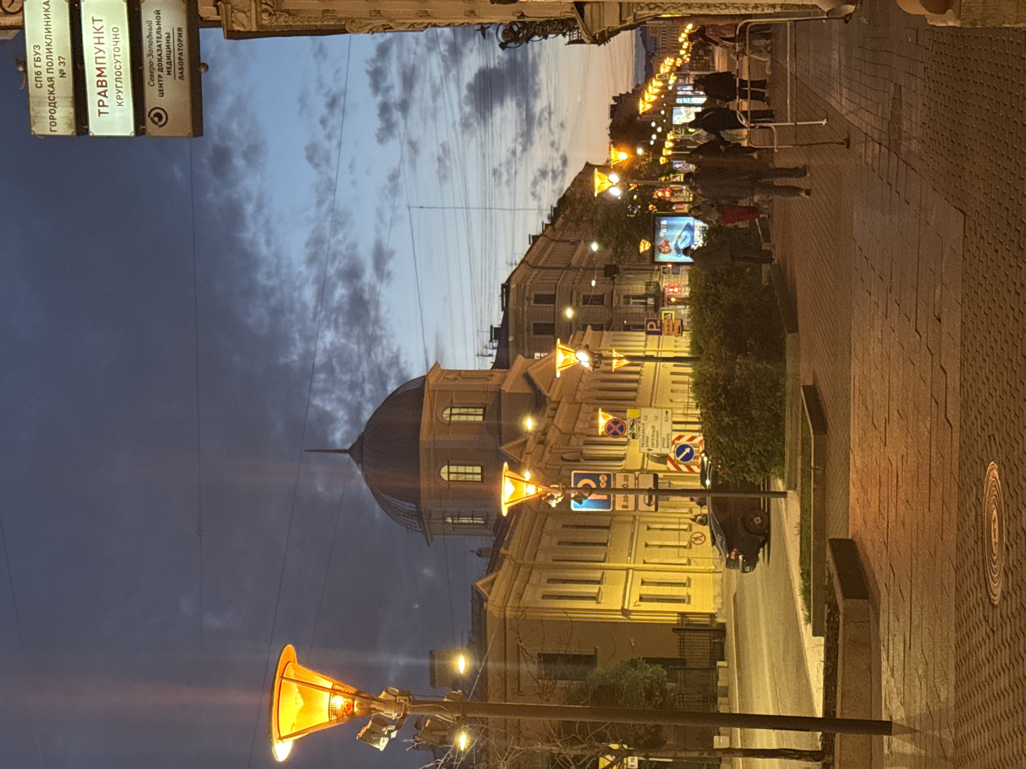 The image depicts a street scene in Saint Petersburg, Russia, at dusk or early evening. The sky is a deep blue with scattered clouds, and the streetlights are illuminated, casting a warm glow on the pavement and buildings. On the left side of the image, a yellow building with a domed roof is visible, likely a historic structure. A streetlamp with a distinctive orange-yellow light stands prominently in the foreground. Across the street, a row of buildings with illuminated windows stretches into the distance, bordered by a line of streetlights. Several signs are visible on the buildings and poles, including one that appears to be a directional sign with Cyrillic text. A few pedestrians are walking along the sidewalk, some of whom are silhouetted against the lights. A dark-colored car is parked on the street. The pavement is made of brick pavers, and there is a manhole cover in the foreground. The overall impression is of a quiet urban evening.