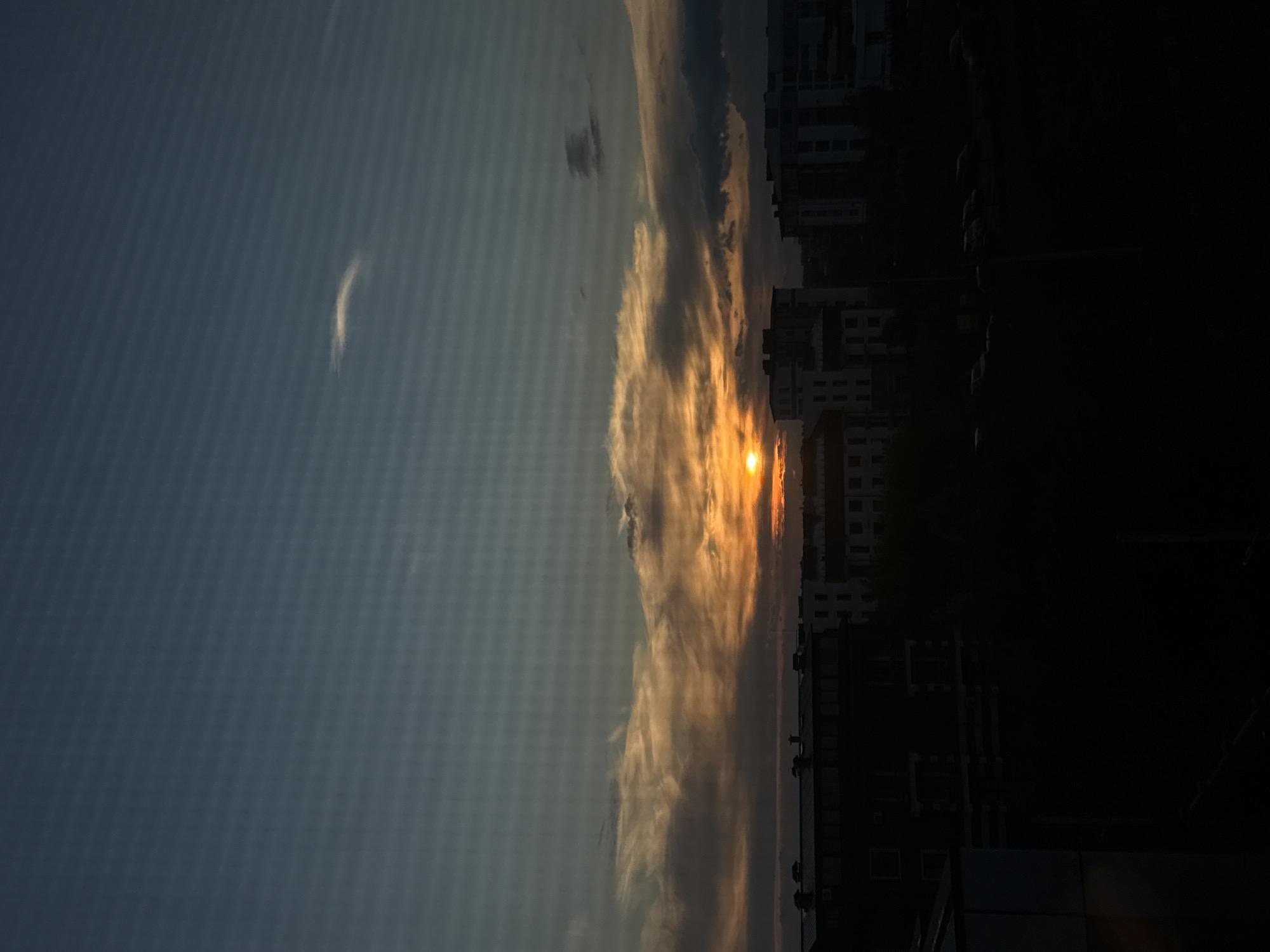 The image captures a sunset over a city skyline. The sun, a bright orange orb, is partially obscured by dramatic clouds, casting warm hues of orange and yellow across the sky. The lower half of the image is dominated by the silhouettes of various buildings, ranging from apartment blocks to what appear to be commercial structures. These buildings are dark against the vibrant sky, with some windows faintly visible. In the foreground, the lower section is almost entirely black, suggesting it is either a very dark area or the perspective is from inside a building looking out. The overall impression is a peaceful, yet somber, urban evening scene.