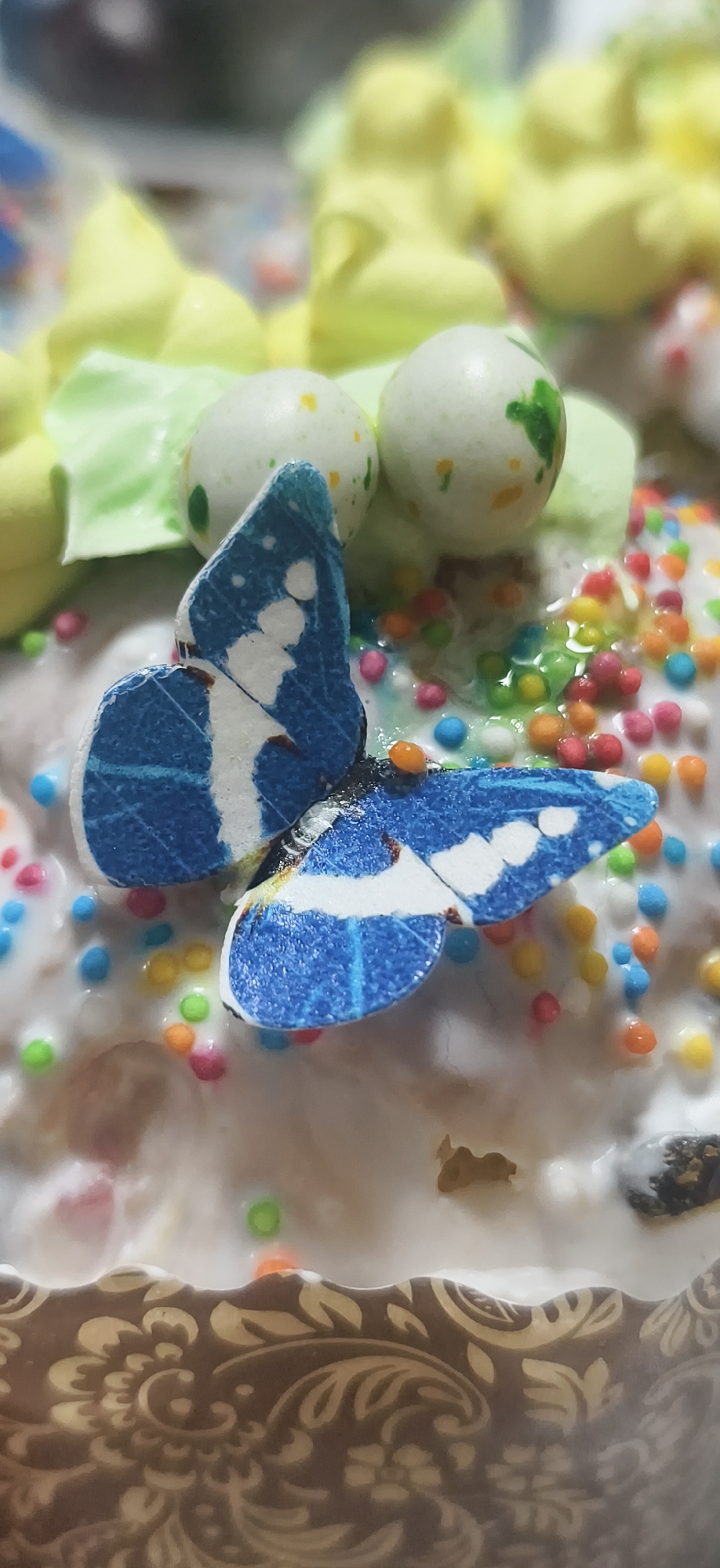 The image shows a close-up of a decorated Easter cake, also known as Kulich. The cake is topped with white icing and colorful sprinkles. A blue and white paper butterfly is placed on top. There are also green and yellow frosting decorations, along with two white, speckled candy eggs. The base of the cake is visible, showing a decorative brown and beige pattern. The location in the city of Фоки, Russia is not directly visible in the image.