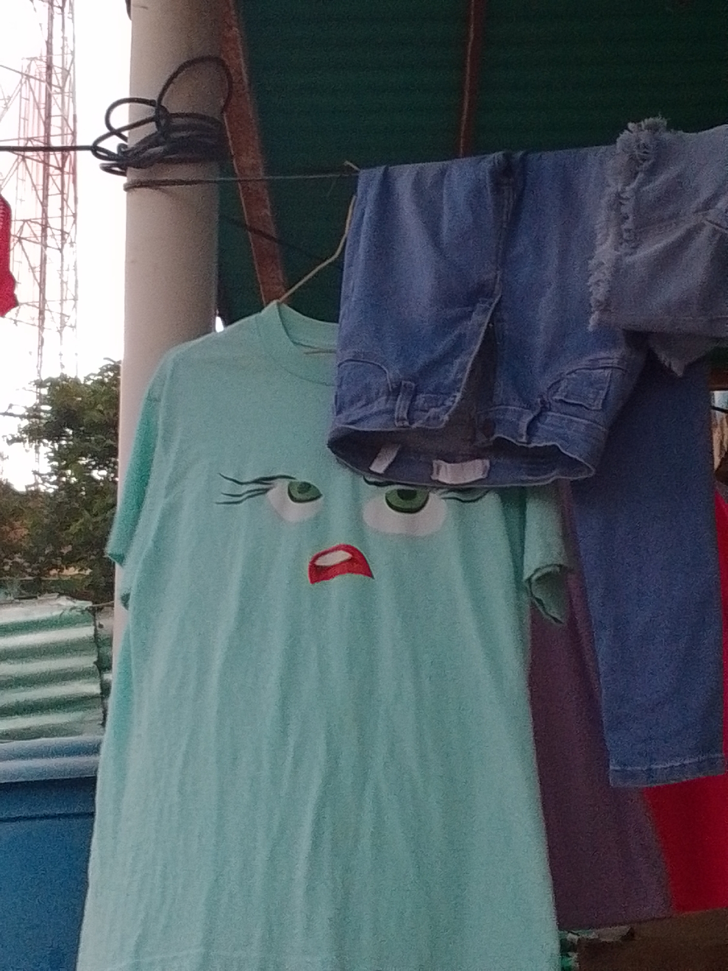 A light turquoise t-shirt with a cartoon face design, featuring large green eyes with eyelashes and an open red mouth, is hanging on a clothesline. The t-shirt is to the left of a pair of blue denim shorts which are also hanging on the clothesline. To the right of the shorts, a dark blue garment, possibly pants, is visible. The clothesline is strung between a white cylindrical pole and what appears to be a green awning. In the background, a portion of a metal tower structure and some green foliage are visible. A red garment is partially visible at the bottom right of the image. The scene is outdoors, and the lighting suggests it is daytime. The location is identified as La Tagua, Venezuela.