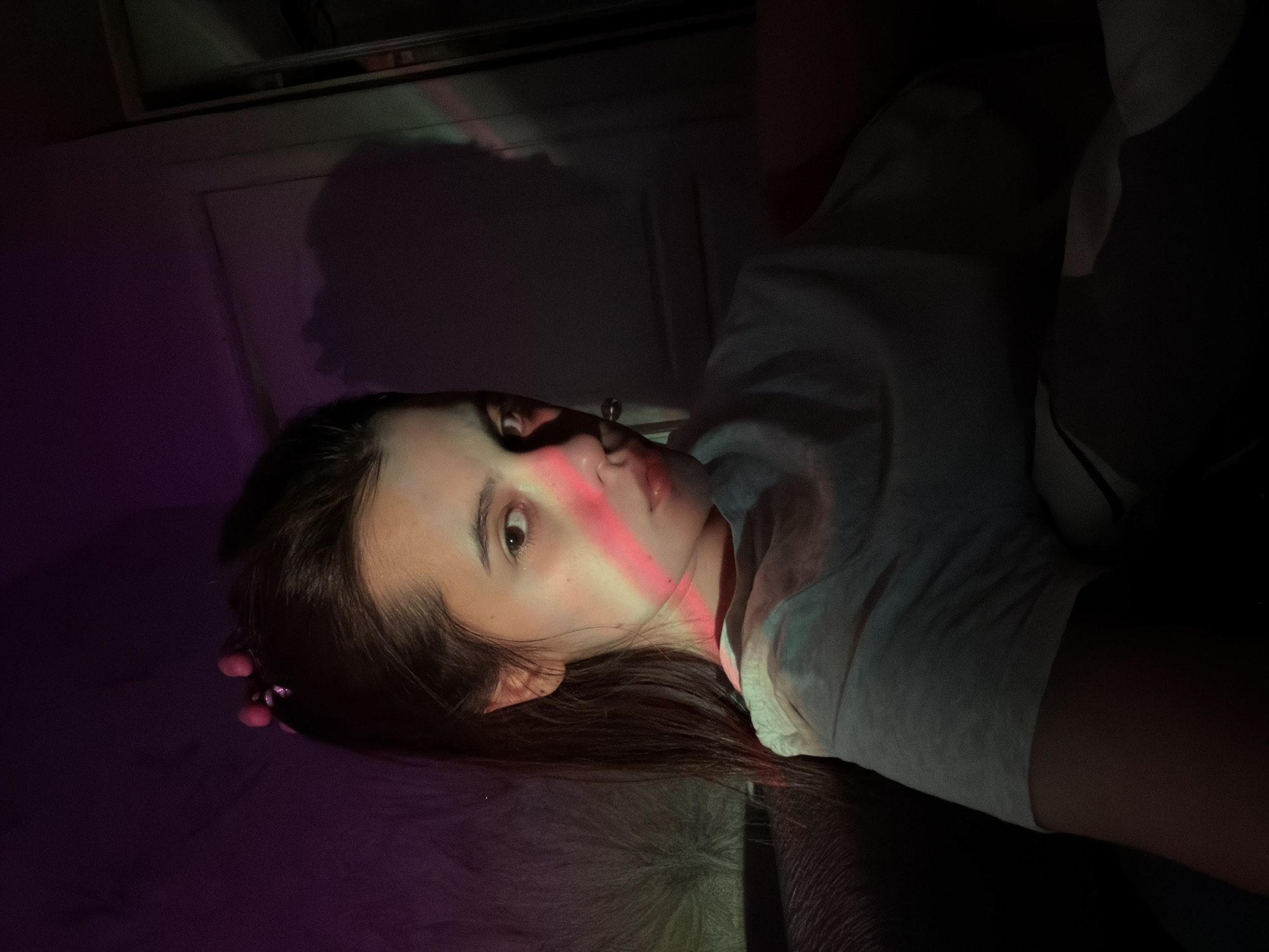 A dimly lit, rotated portrait captures a young person, likely female, lying down indoors. Her dark hair spreads out around her head as she looks directly towards the camera with a neutral, slightly serious expression. A prominent red-pink beam of light sharply cuts across her face, illuminating her right cheek, eye, and lips, and creating a striking contrast with the darker areas of the image.
She is wearing a light-colored (possibly white or grey) top, visible on her torso and arm. On her left ear, a decorative earring with small, round, pink elements is visible. The background is predominantly dark, suggesting an indoor setting with minimal ambient light. A faint, multi-colored light projection, resembling a small rainbow, is visible on a dark wall or door in the upper left background. Below her, a textured, dark surface, possibly a blanket or furniture, is visible. On the dark wall behind her, a faint shadow of a profile, possibly her own, is cast.
The overall atmosphere is moody and artful, created by the interplay of shadow and the artificial colored light. The image appears to be taken at night or in a room with controlled lighting. There is no visible text or specific location cues within the image itself, though the context indicates the location is Chaikovsky, Russia.