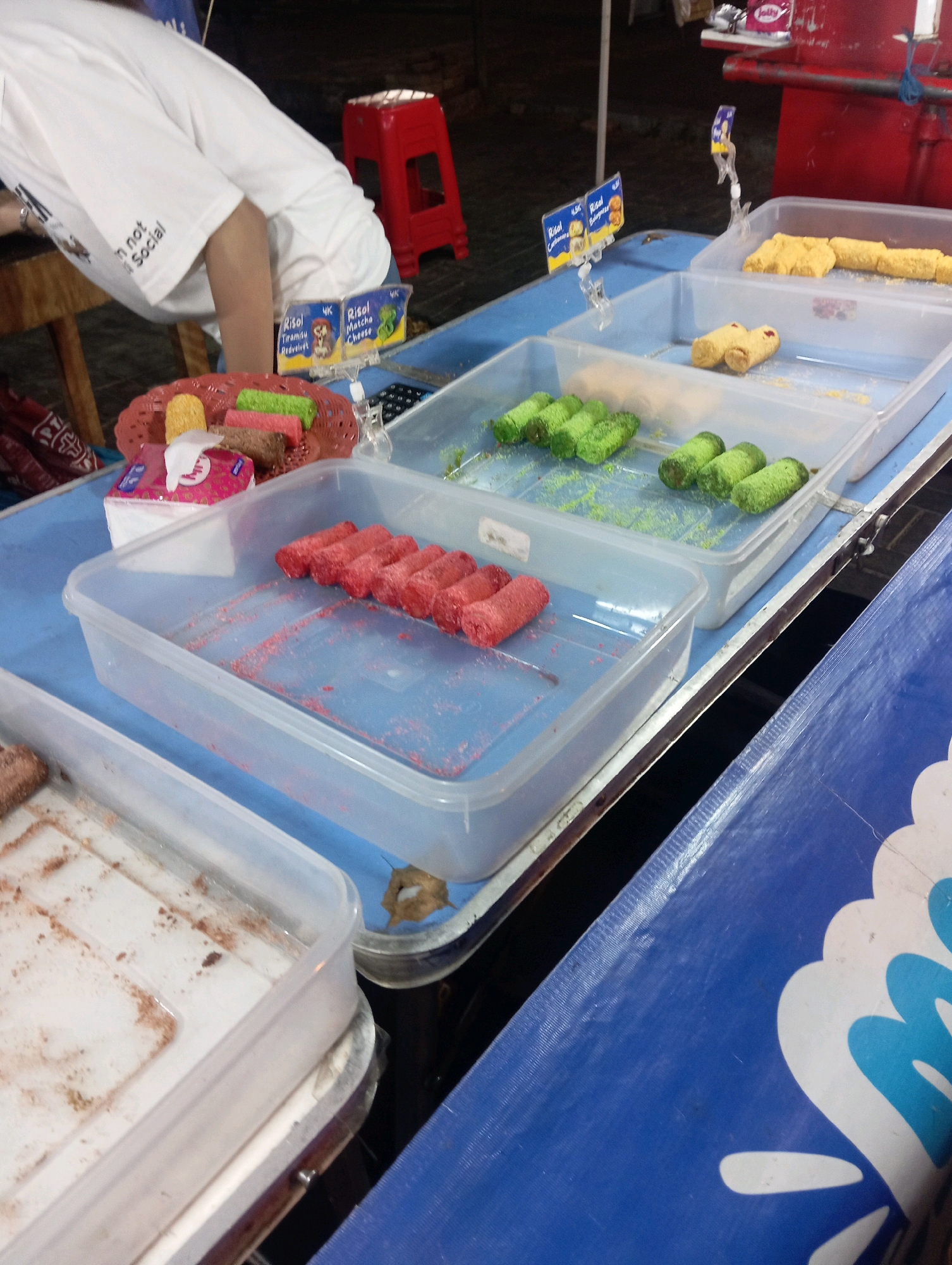 In Magelang, Indonesia, a street-side food stall displays a variety of snacks. A person wearing a white t-shirt with the text "I'm not social" is partially visible, leaning over a table. On the table, there are clear plastic containers filled with food items. One container holds several green cylindrical snacks, another has red cylindrical snacks, and a third has yellow, elongated snacks. A small red plastic stool is visible in the background. There are also product labels, one with "Risol Tiramisu Redvelvet" and another with "Risol Matcha Cheese". The scene appears to be an outdoor setting, possibly a night market or a temporary stall.