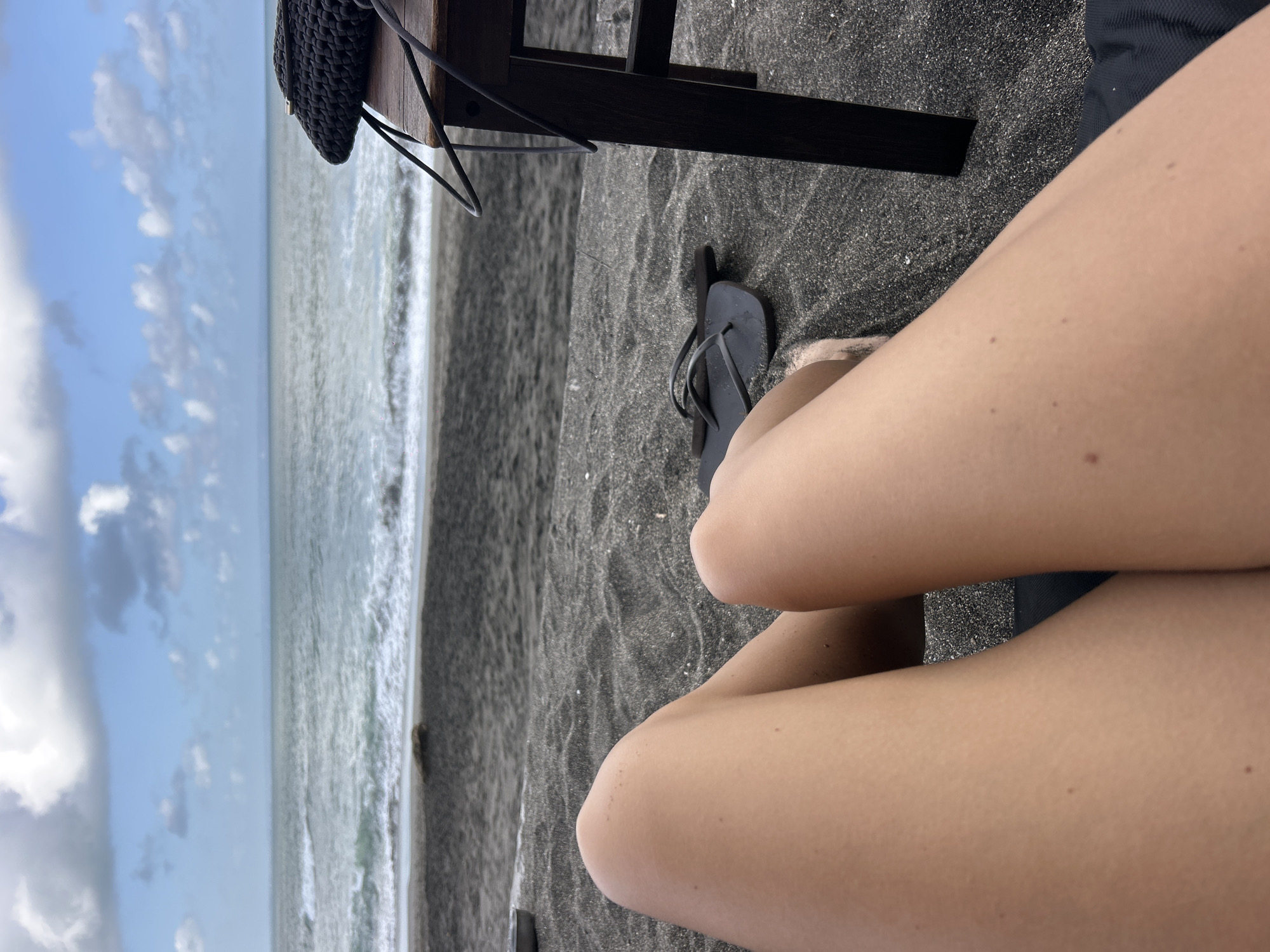 The image shows a person sitting on a beach with dark-colored sand, possibly volcanic sand, in Mengwi, Indonesia. The person's legs are visible in the foreground, positioned in a crossed or relaxed manner. A pair of dark-colored flip-flops is on the sand. A table is partially visible behind the legs, with a woven bag hanging from it. The ocean is visible in the background, with waves approaching the shore and a partly cloudy sky above.