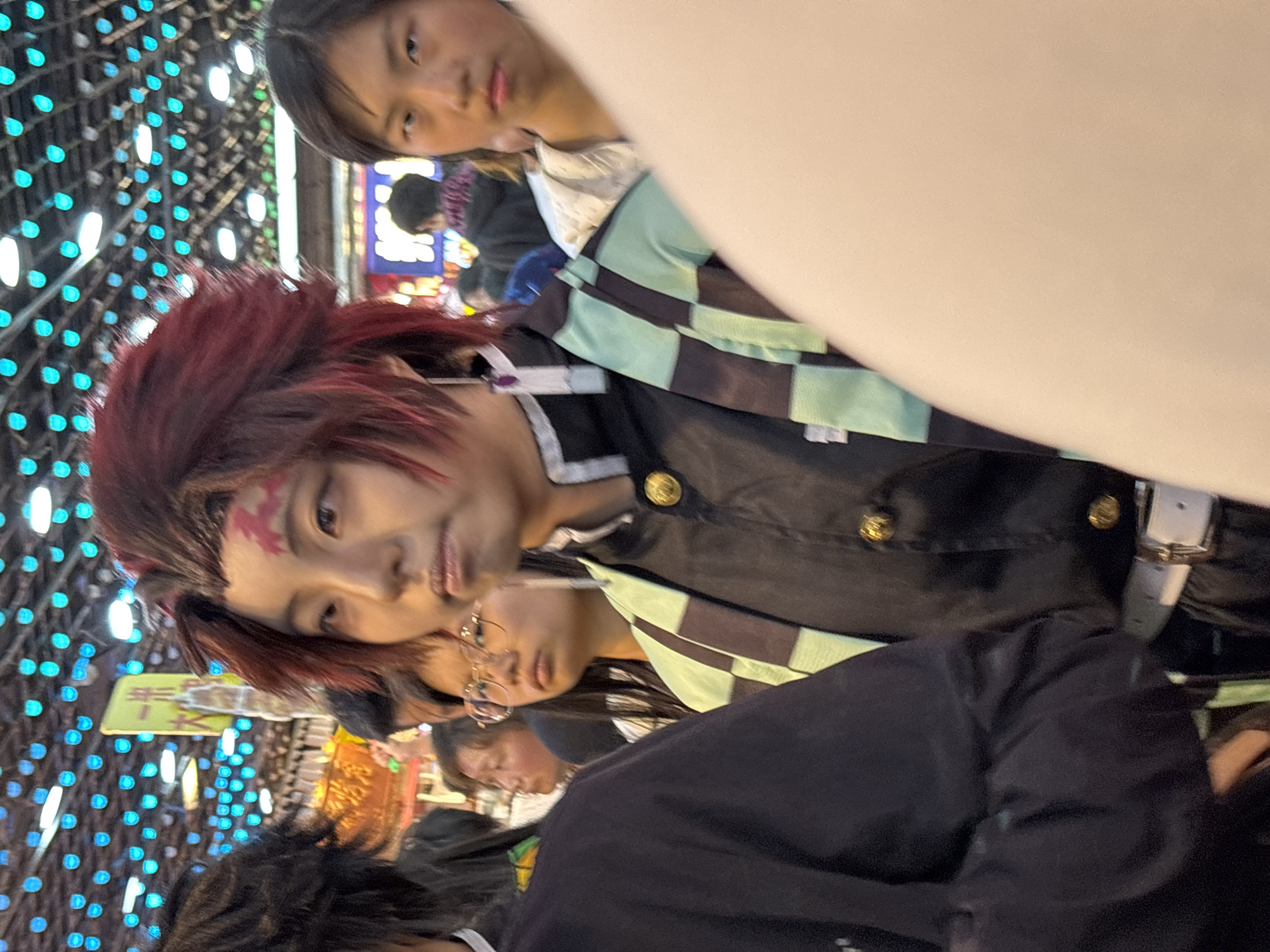 In a brightly lit, crowded urban environment, several people are visible, likely at a gathering or event. The primary subject is dressed in a distinctive costume with red hair and facial markings, suggesting cosplay or a character portrayal. They are wearing a black jacket with gold buttons, a patterned scarf, and a white belt. Another person stands close by, with a portion of their face and dark hair visible. Below them, another individual wears glasses and a dark shirt. Behind them, a person with dark hair and a dark jacket is partially visible. Strings of small, bright lights create a backdrop of blue and white dots, contributing to the festive atmosphere. The scene suggests a public event or gathering, possibly a convention or festival, in the city of Unknown, Unknown.